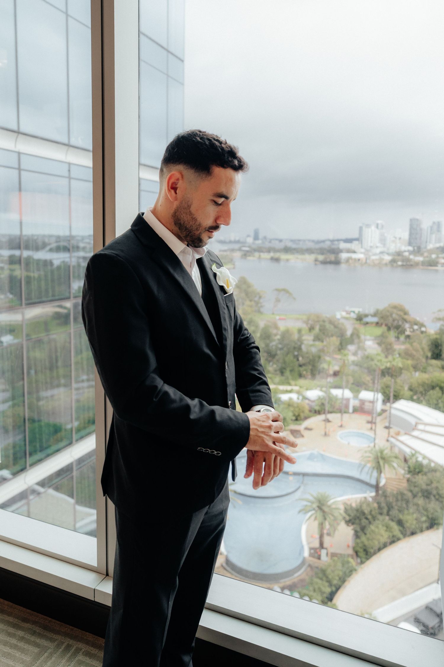 Groom in black suit with white boutonniere checking his watch by a high-rise window with city views.