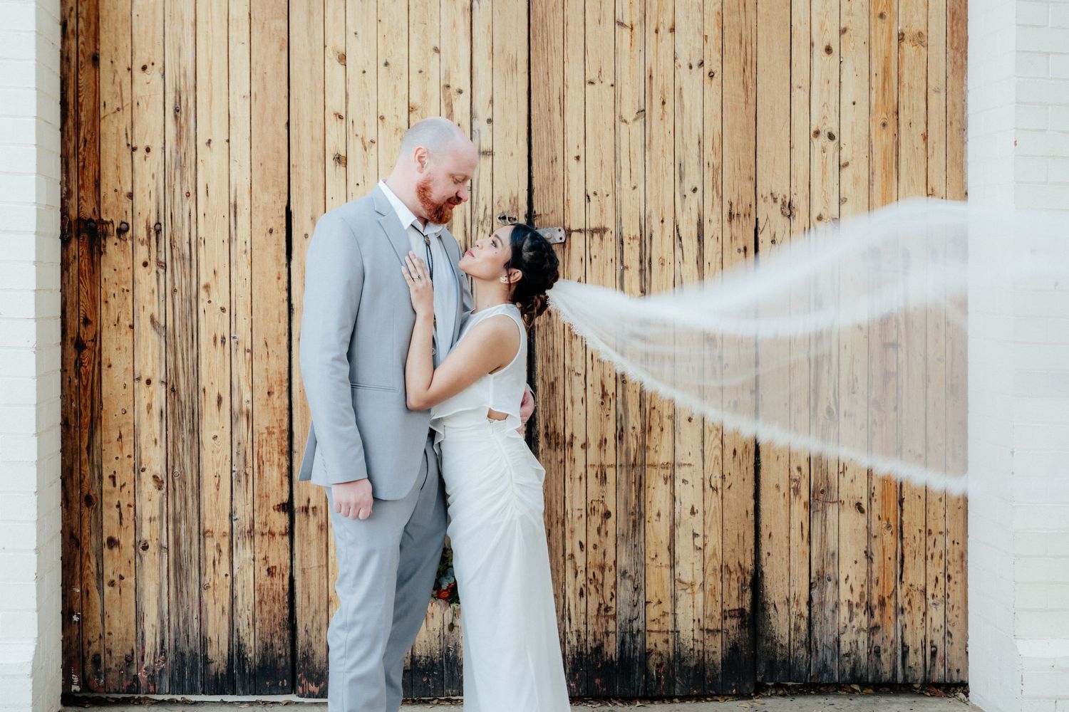 Bride and groom gazing at each other with veil flowing beside rustic wooden barn doors.