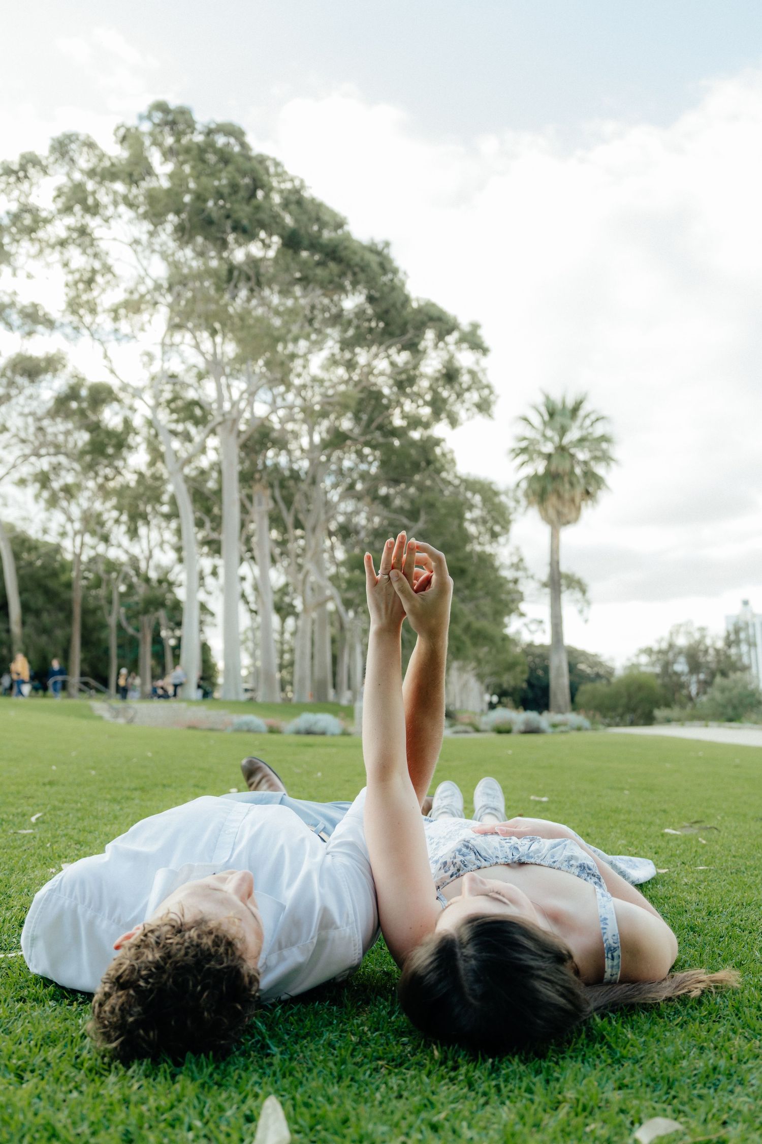 Couple lying on green grass in a park holding hands raised up toward the sky.