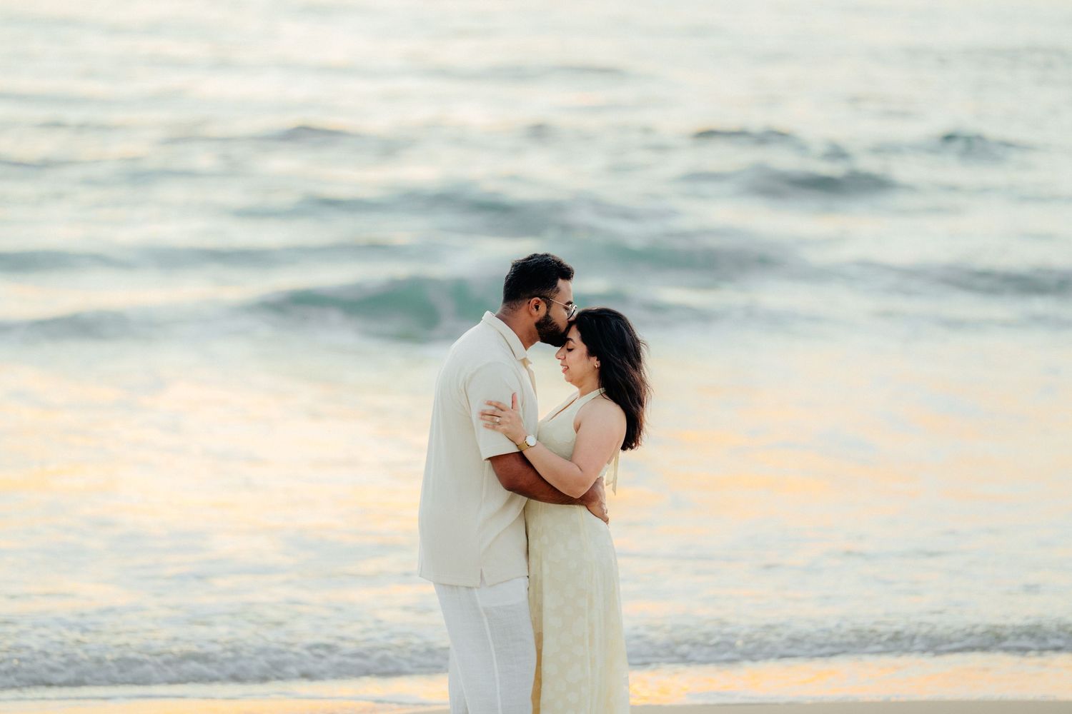 Couple embracing intimately on the beach with golden sunset ocean waves behind them.