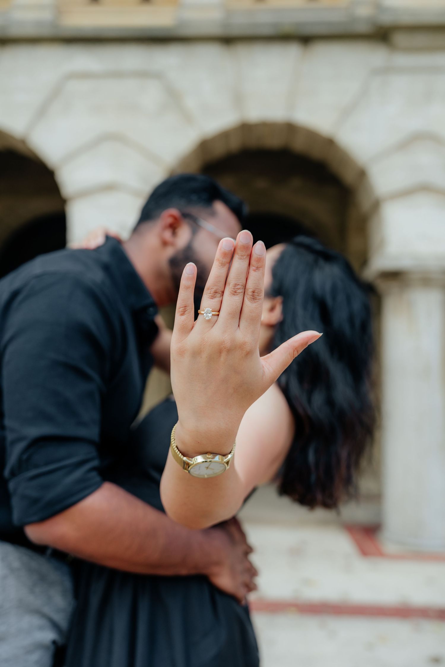 Couple kissing while showing off a gold engagement ring in front of an ornate stone building.