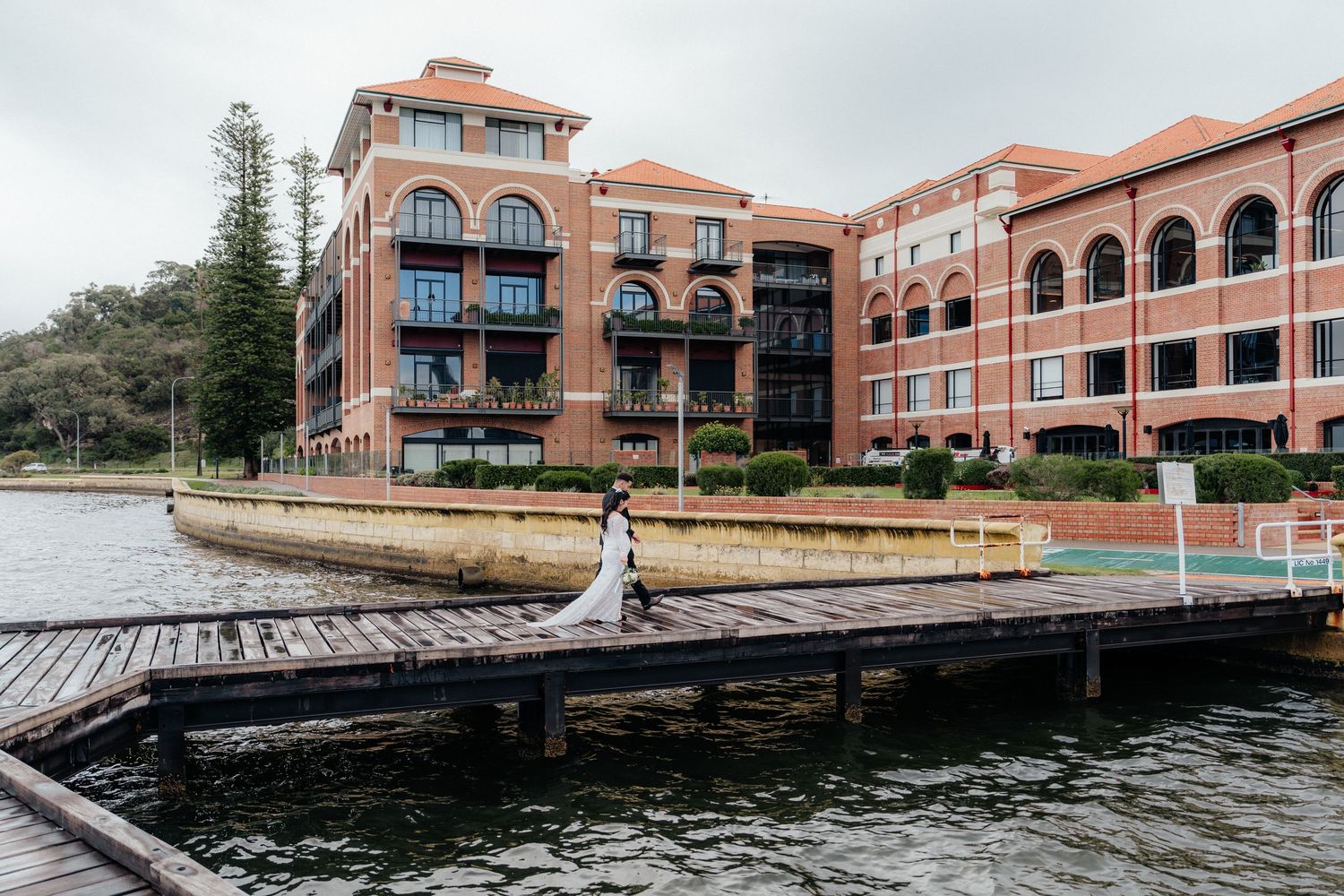 Bride in white wedding gown poses on waterfront dock with elegant brick hotel building in background.