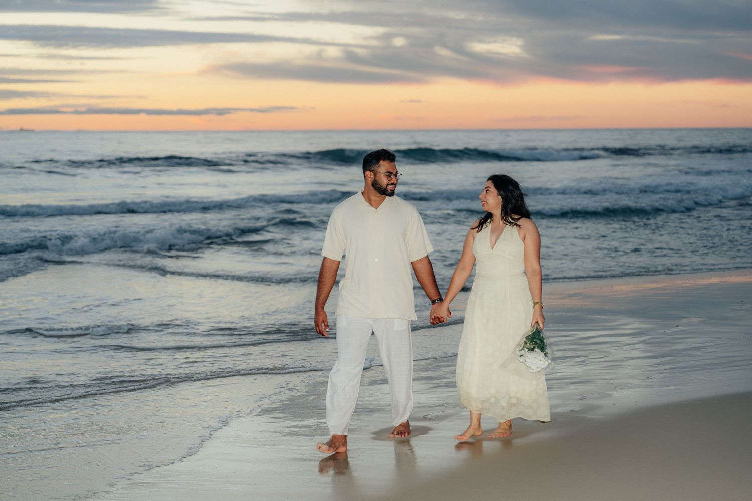 Couple in white holding hands on beach at sunset with ocean waves behind them.