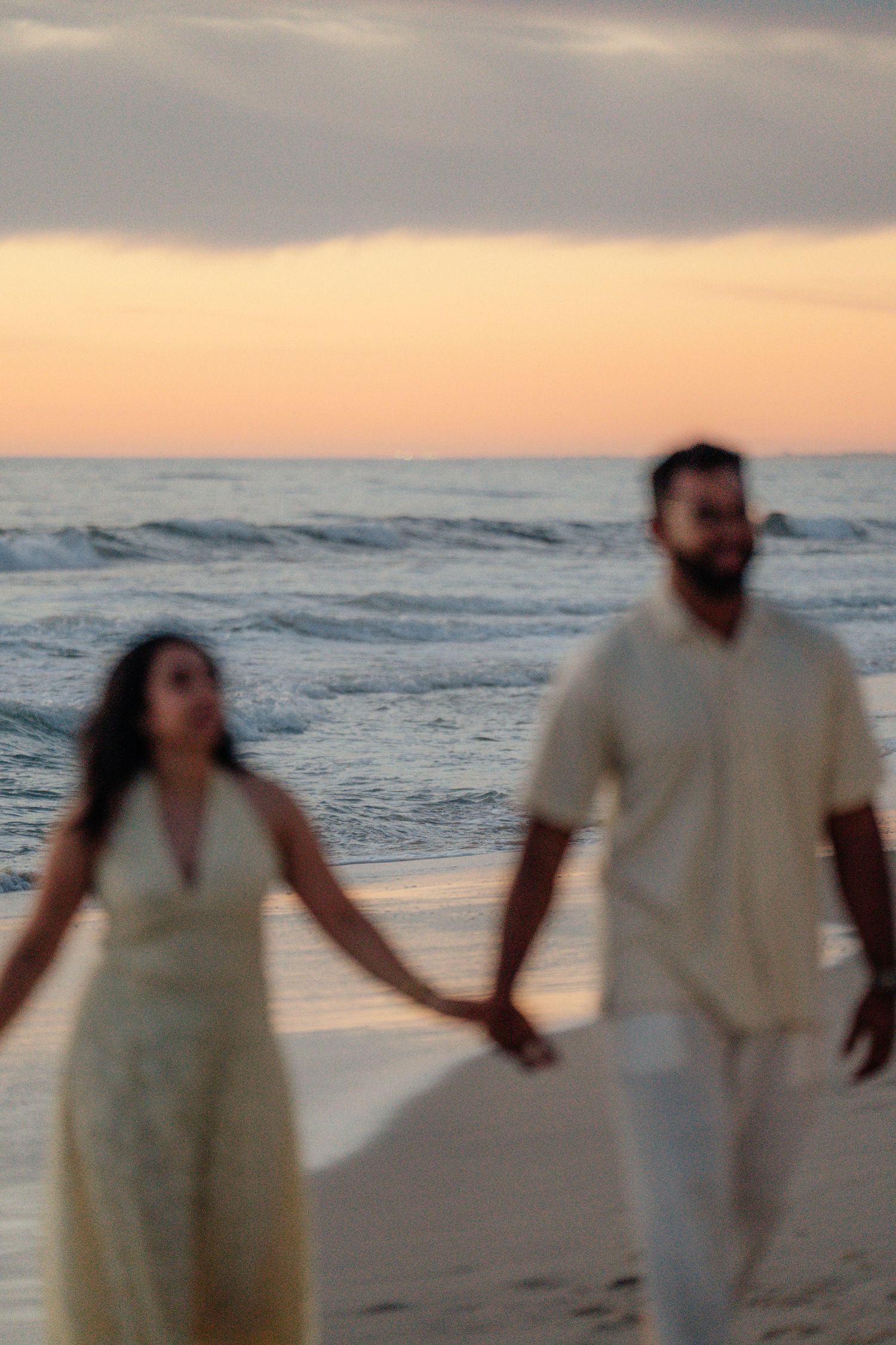 Soft focus couple holding hands walking along sandy beach with pastel sunset sky.