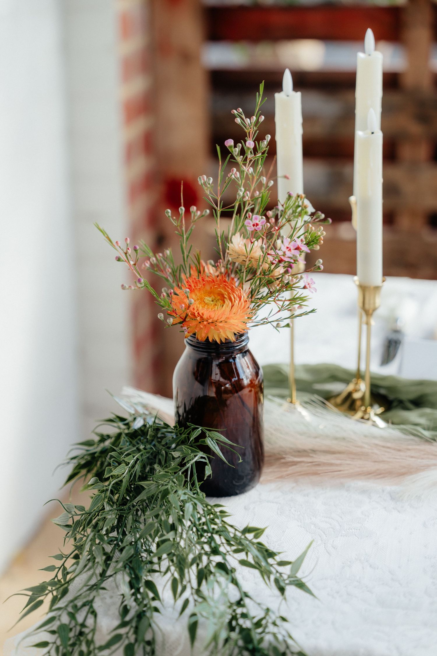Rustic wedding table decor with amber vase of wildflowers, white taper candles, and eucalyptus greenery runner.