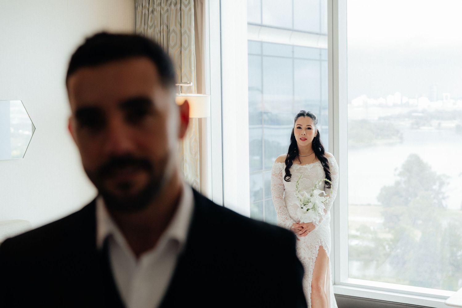 Bride in white lace gown stands by window as groom in black suit stands in foreground during wedding preparations.