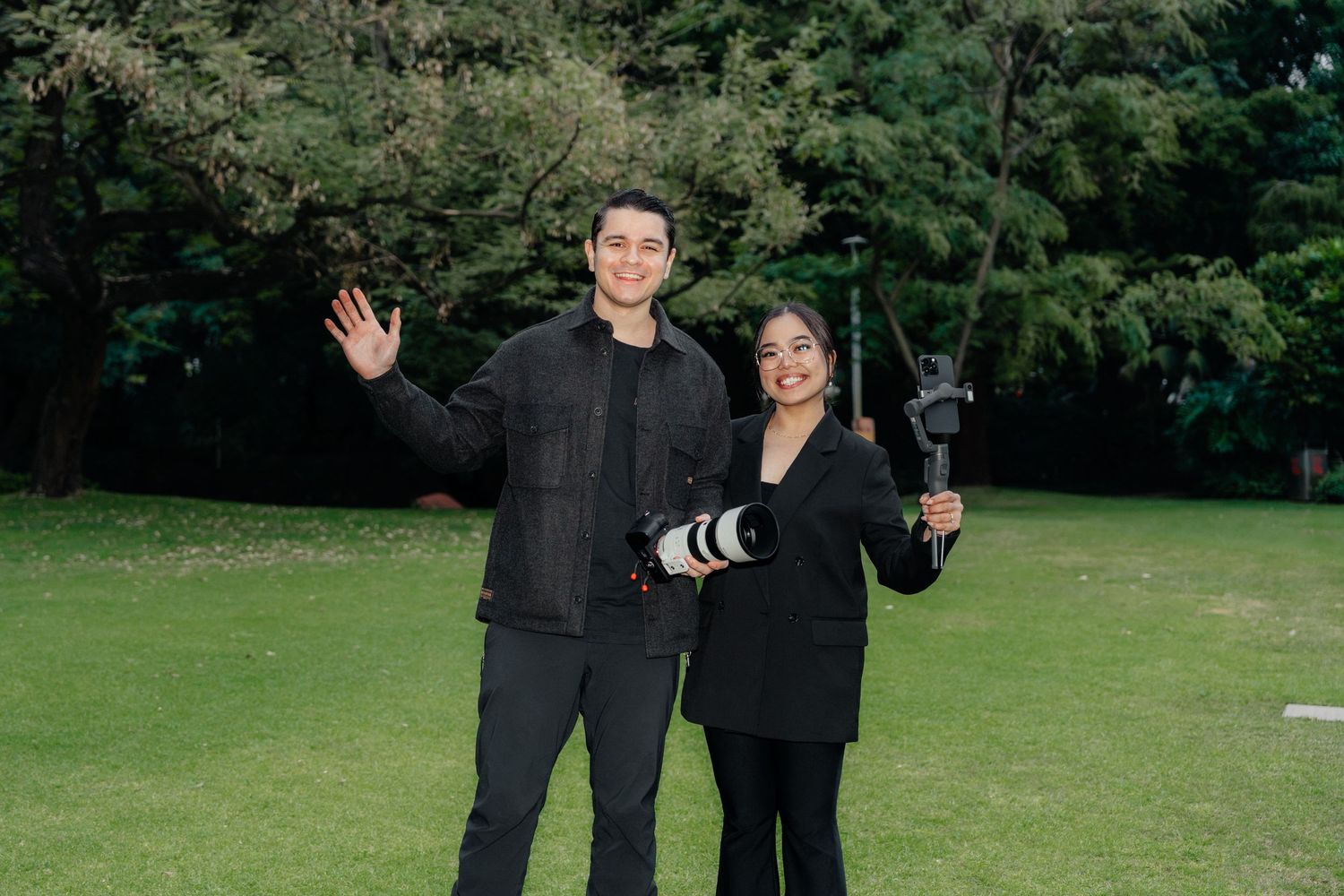 Two photographers smiling outdoors holding a camera and gimbal stabilizer in a green park.