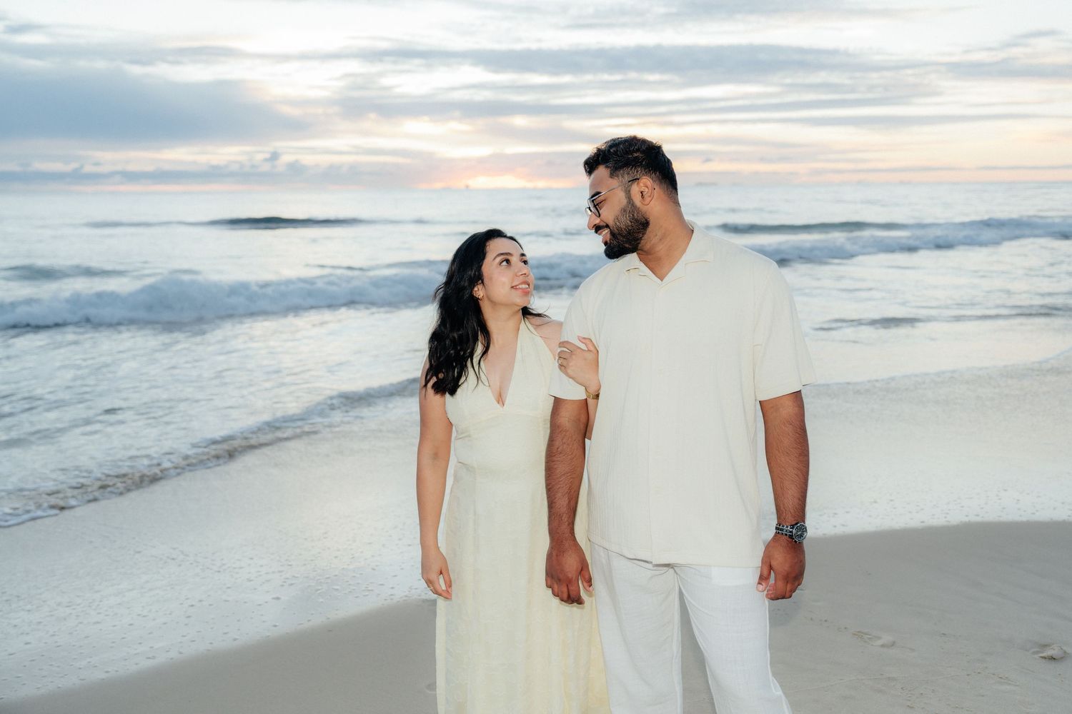 Happy couple in white outfits walking hand in hand on a sandy beach at sunset.