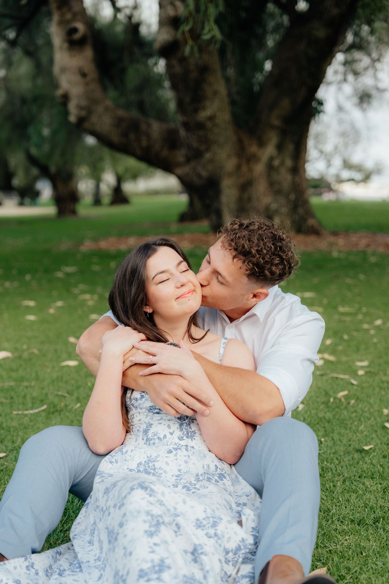 Man kissing woman on cheek while embracing her from behind in a lush park setting.