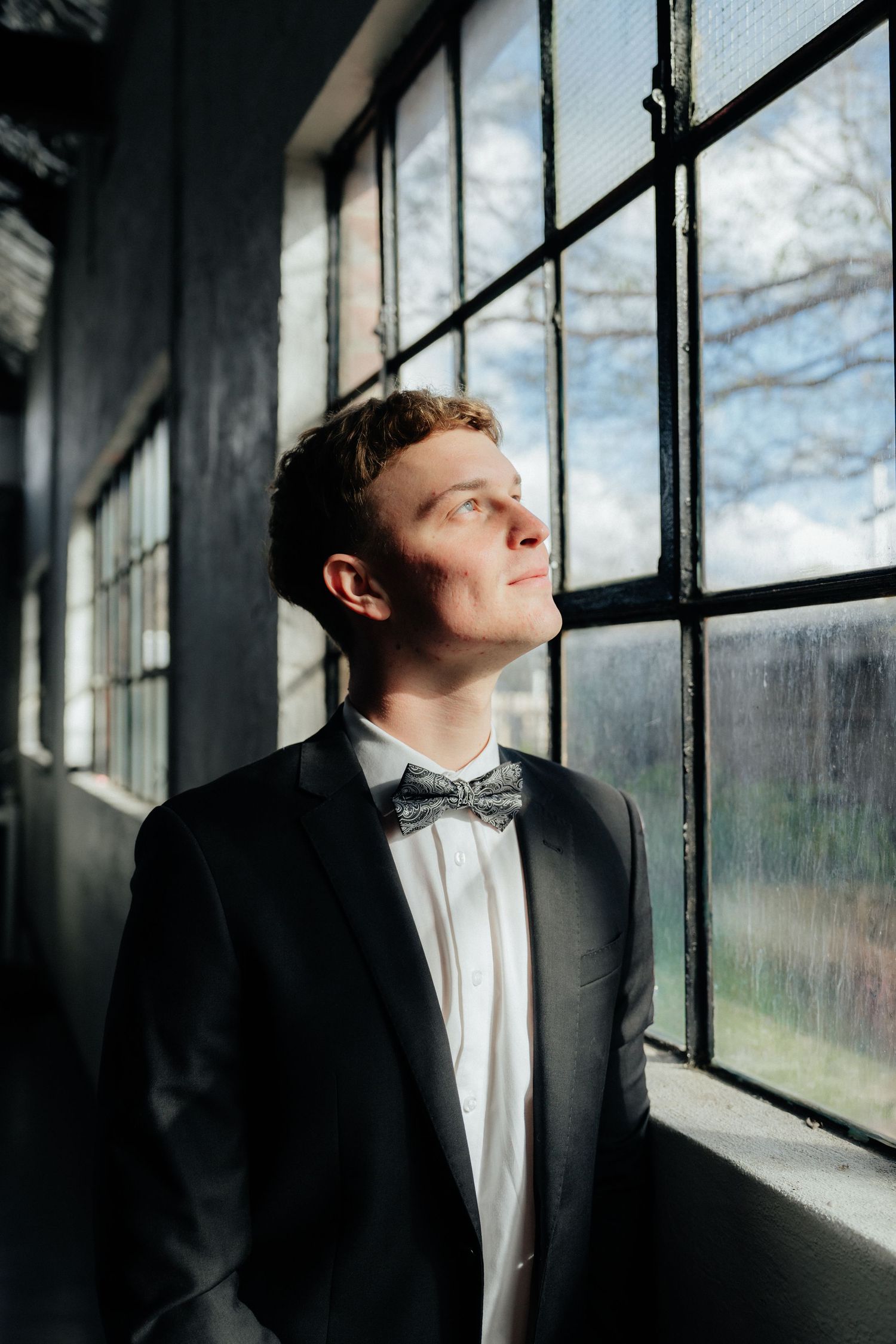 Handsome groom in black tuxedo and bow tie gazing upward by industrial warehouse windows.
