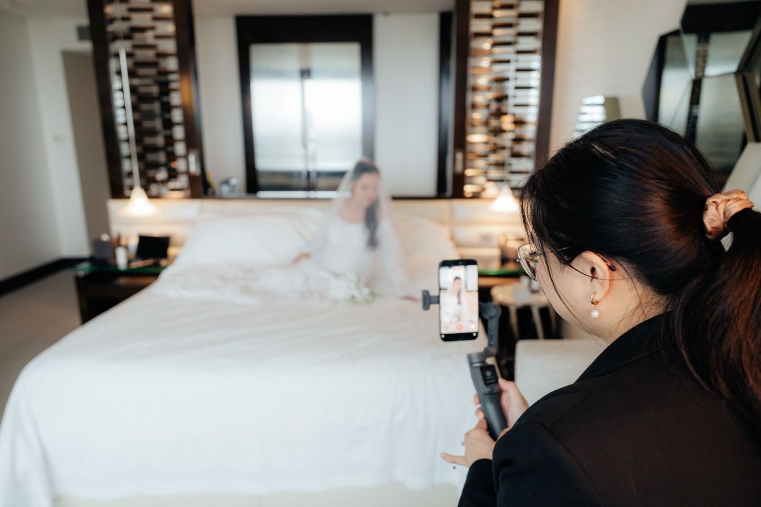 Bridesmaid photographs bride posing on white hotel bed in bridal preparation scene.
