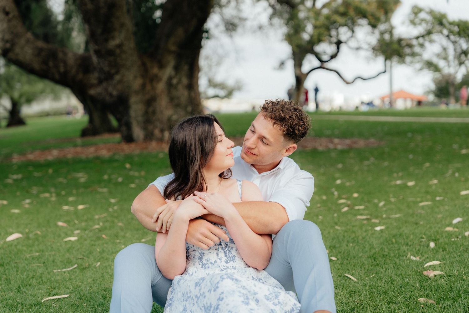 Couple sitting on grass embracing under large tree in romantic outdoor engagement photo.