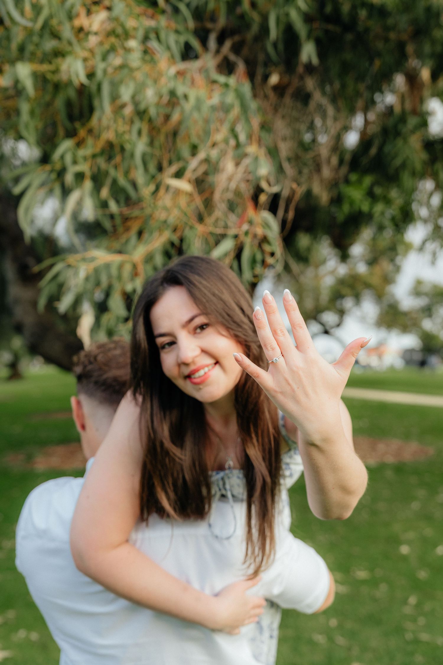 Happy woman showing off engagement ring while being embraced by her partner in a park.