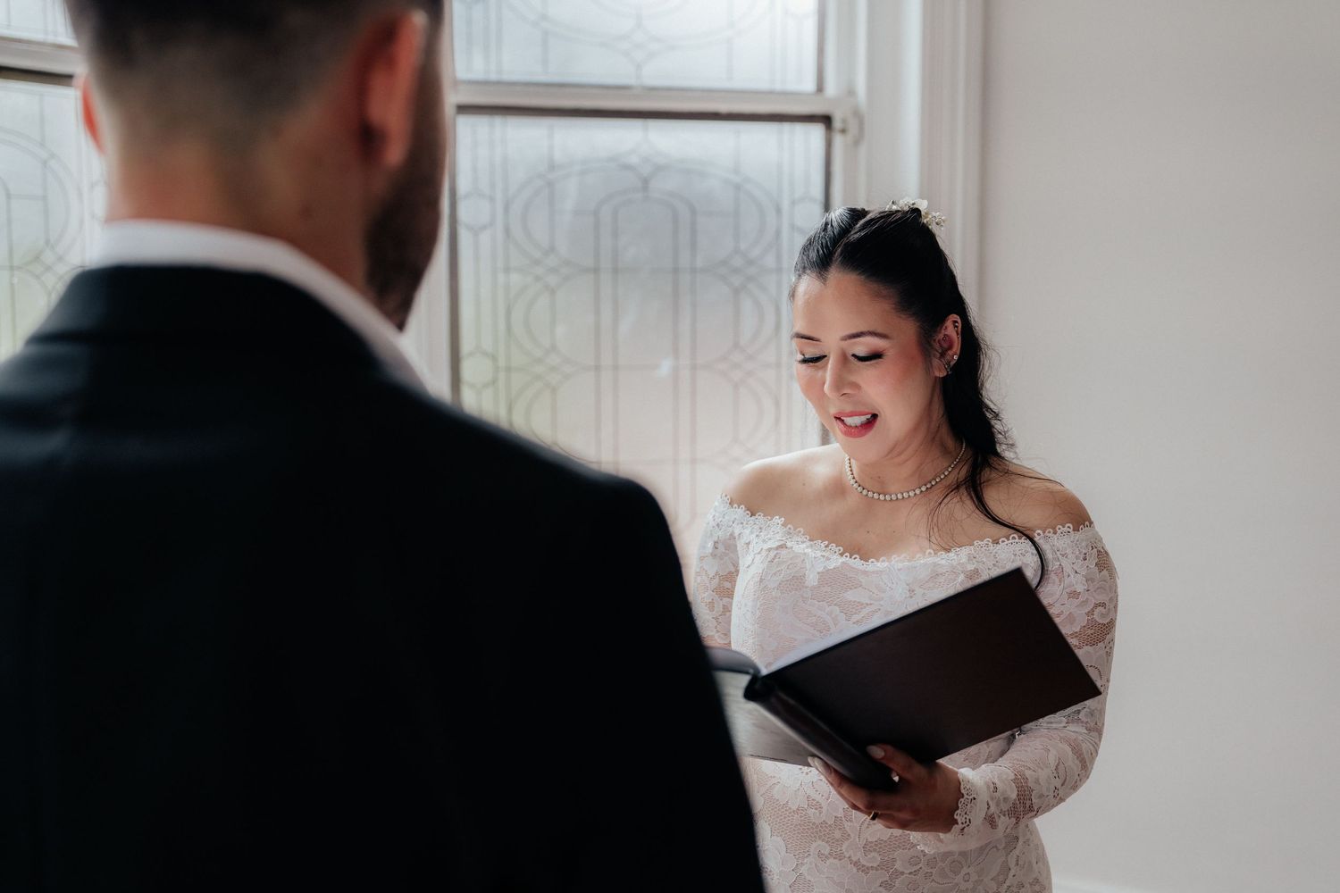 Bride in lace off-shoulder wedding dress reads vows from a black folder, smiling, with groom in black suit in foreground.