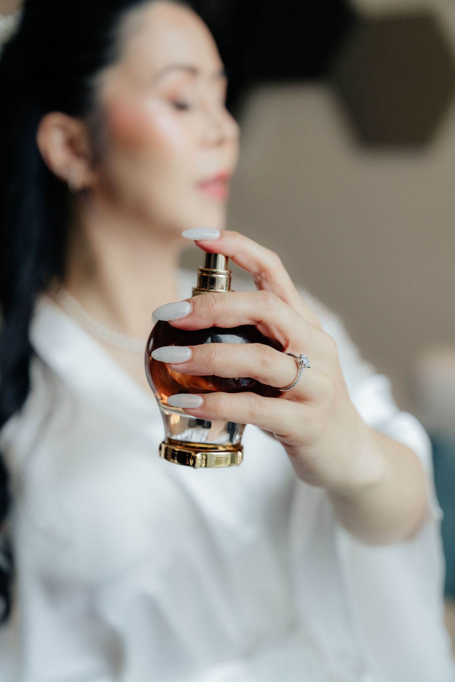 Bride in white robe holding luxury perfume bottle while wearing diamond engagement ring during wedding prep.