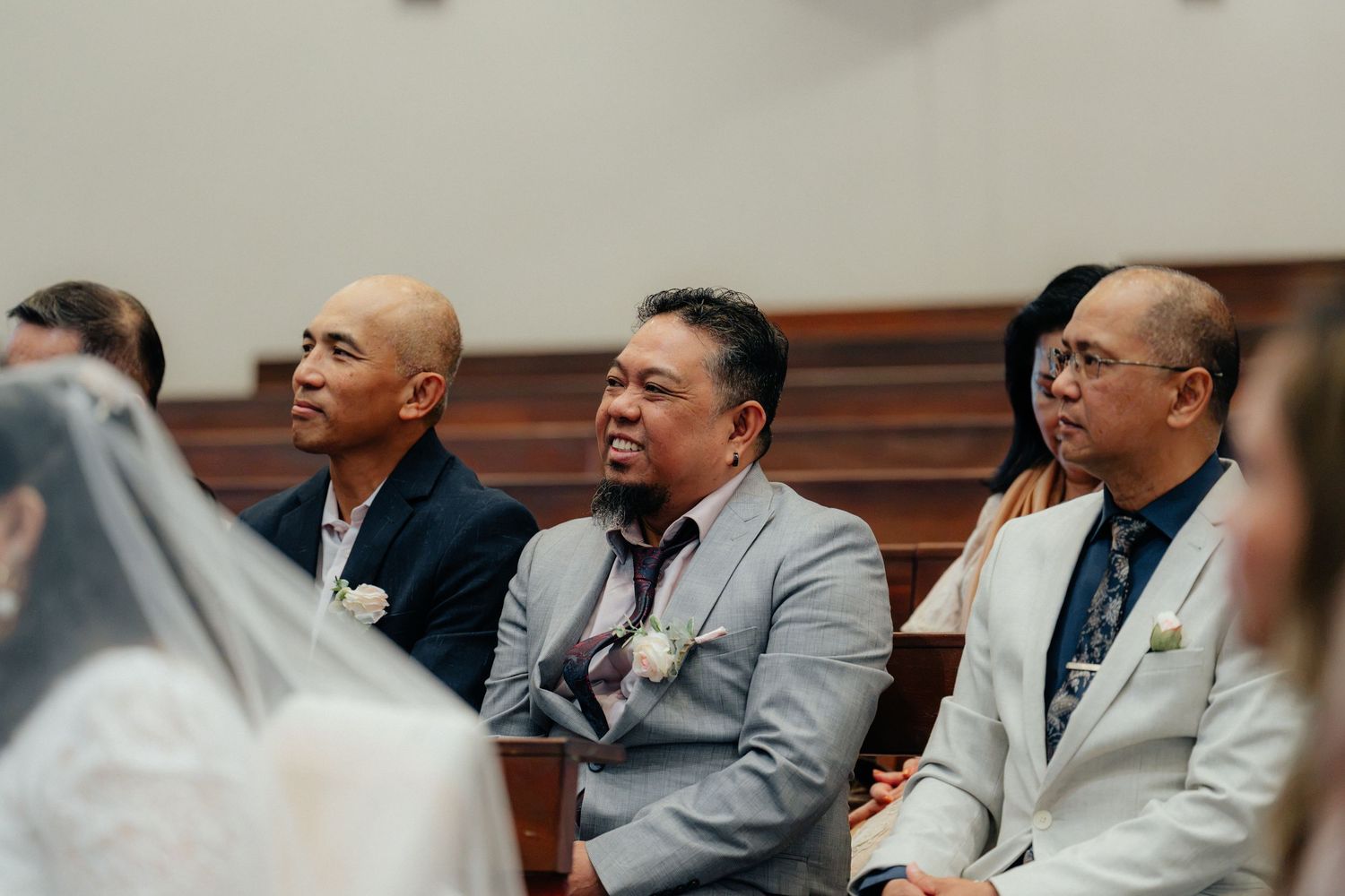 Three male wedding guests seated in church pews smiling and watching the wedding ceremony proceedings.