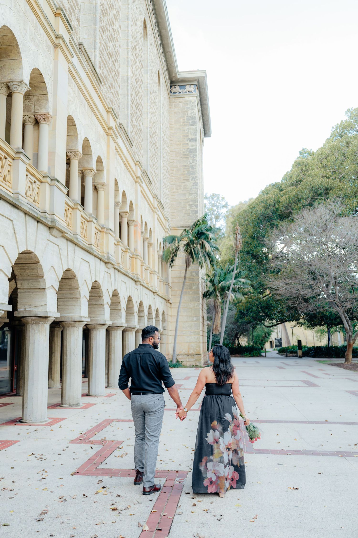 Couple holding hands walking past a grand stone building with arched colonnades and palm trees.