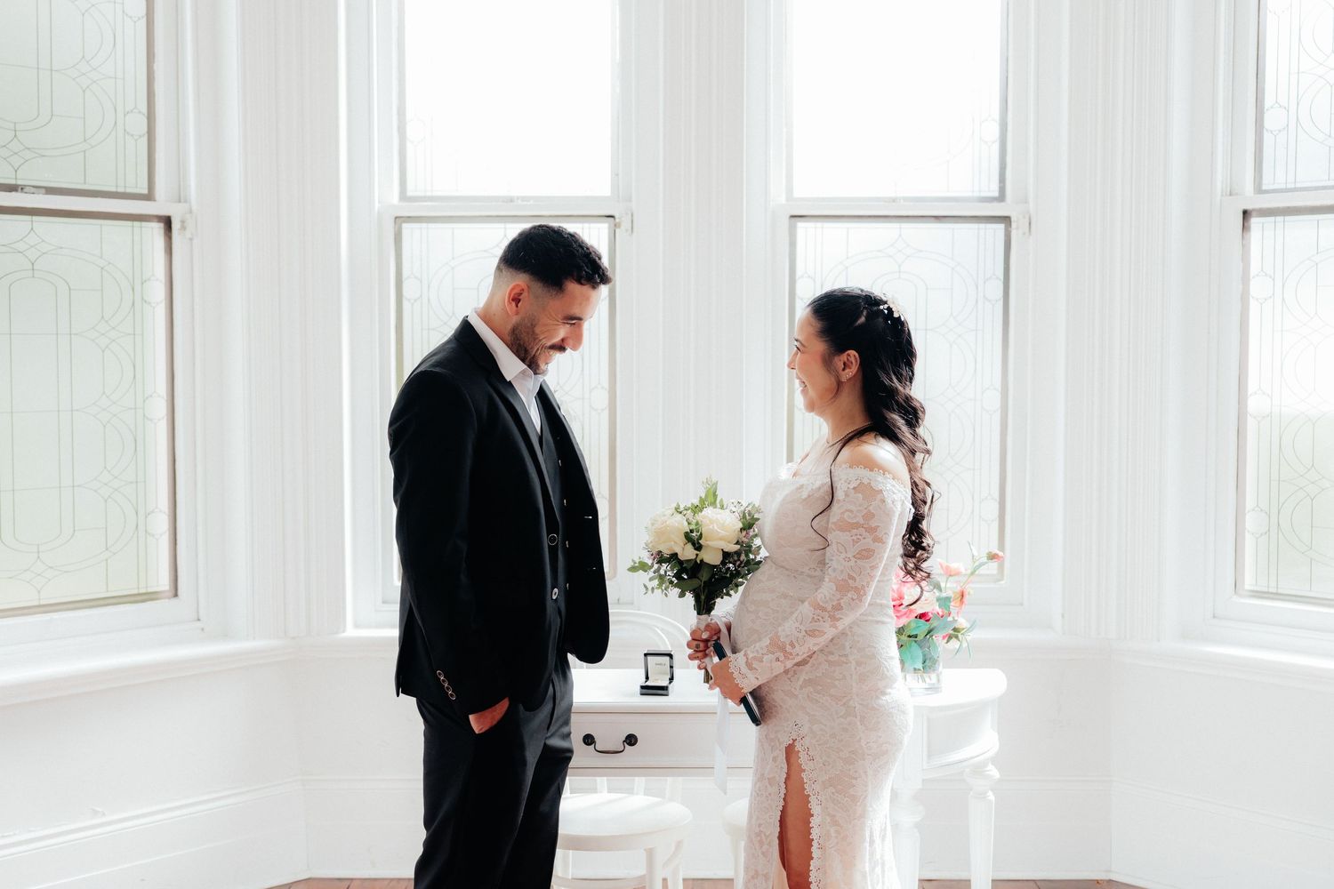 Bride and groom share an intimate first look moment by bright windows, bride holding white bouquet.