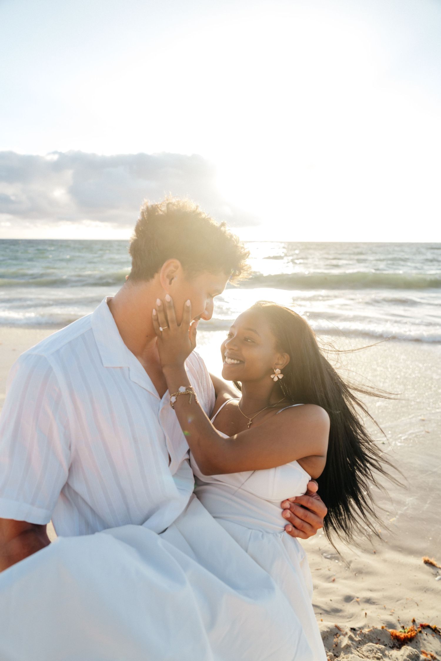 Couple sharing a romantic moment on a sunlit beach, smiling at each other in white outfits.