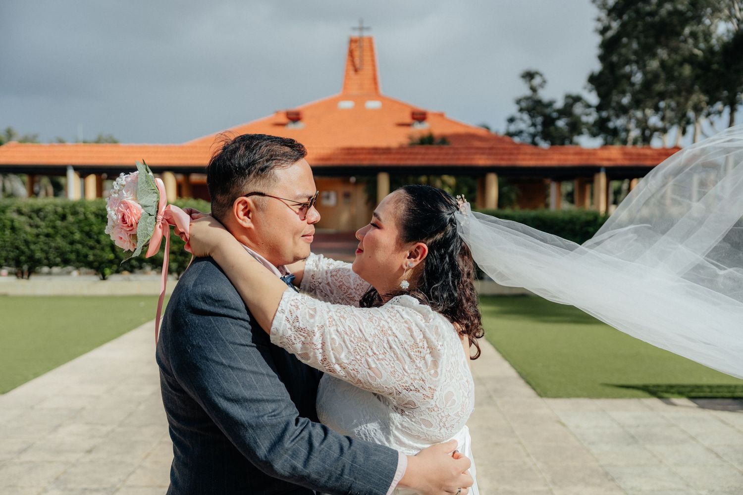 A bride and groom embrace lovingly outdoors with the bride's veil flowing in the breeze.