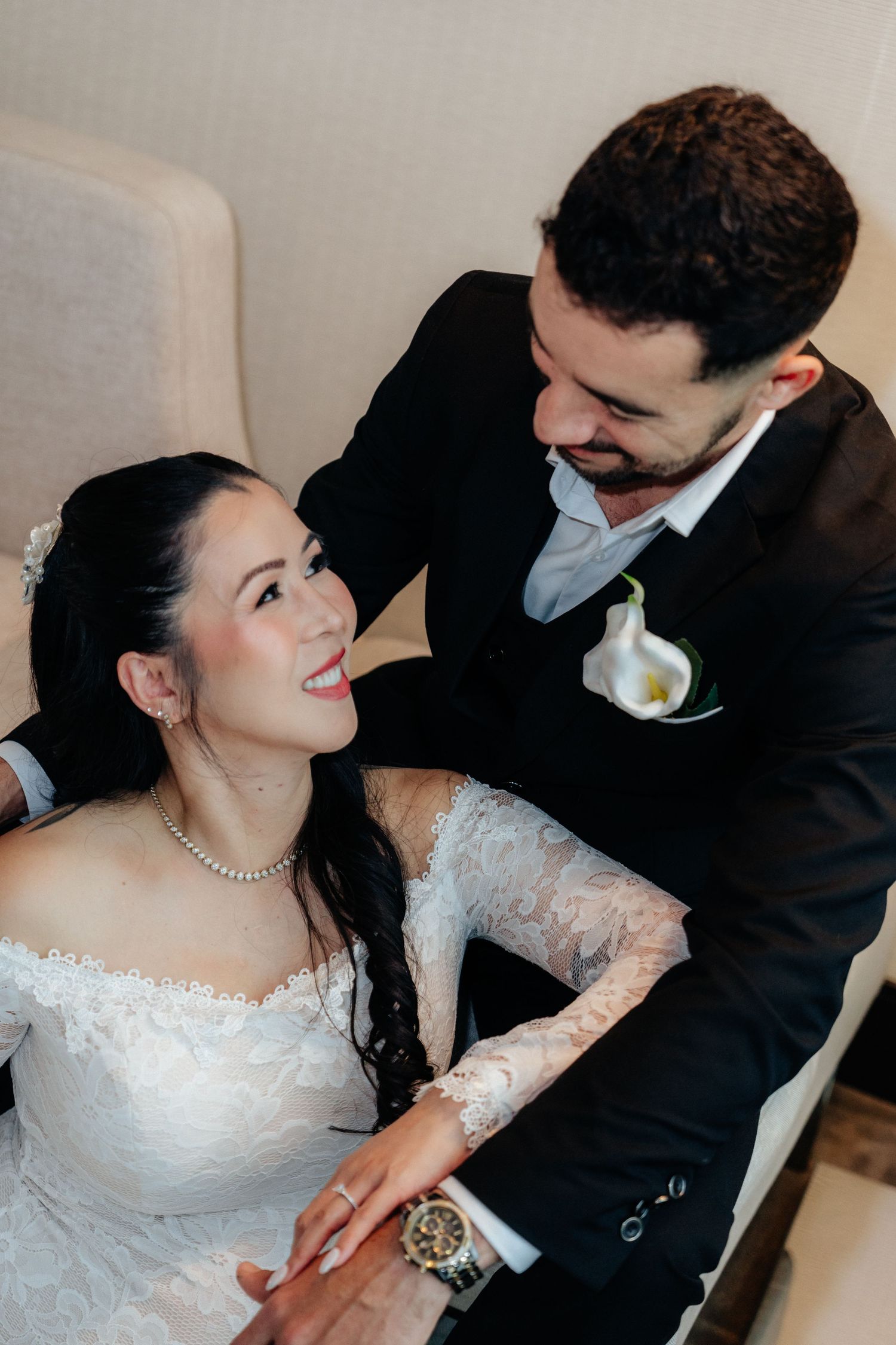 Bride in lace gown smiles up at groom in black suit during tender wedding moment.