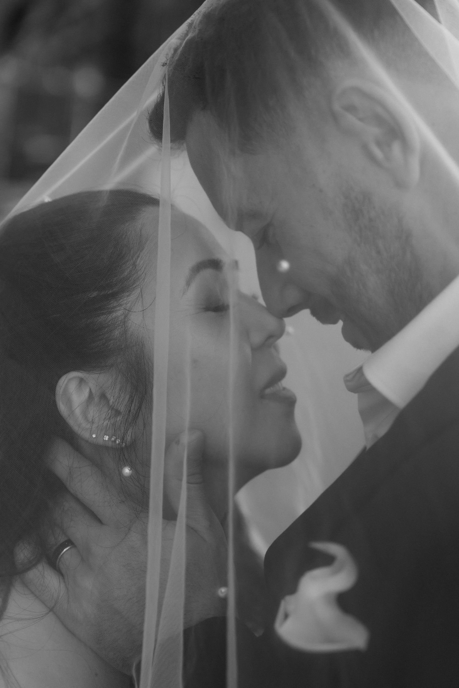 Romantic black and white photo of bride and groom touching foreheads under wedding veil in tender moment.