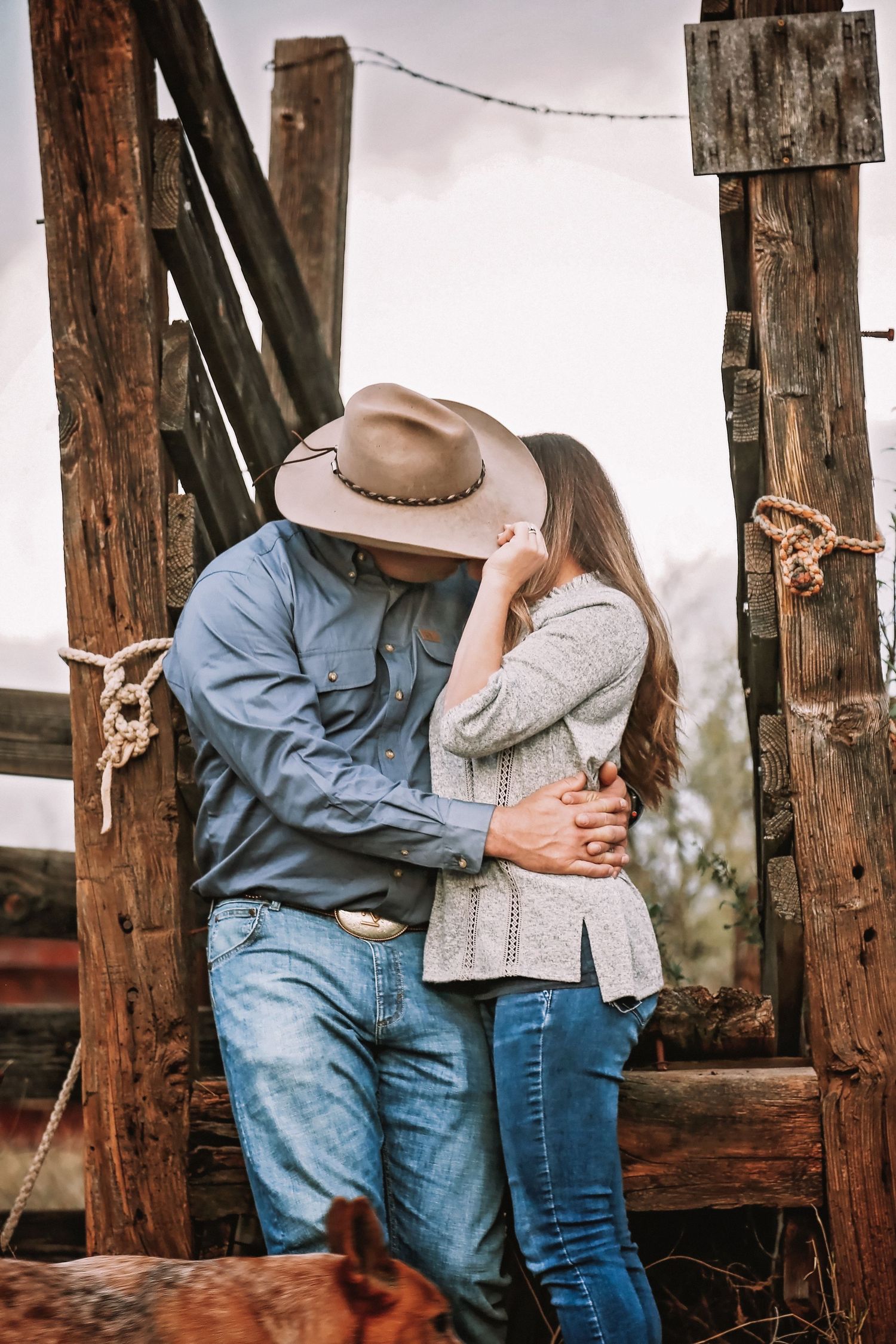 Cowboy in a tan hat and denim shirt kisses a woman in a cardigan and jeans beside a rustic wooden fence post.