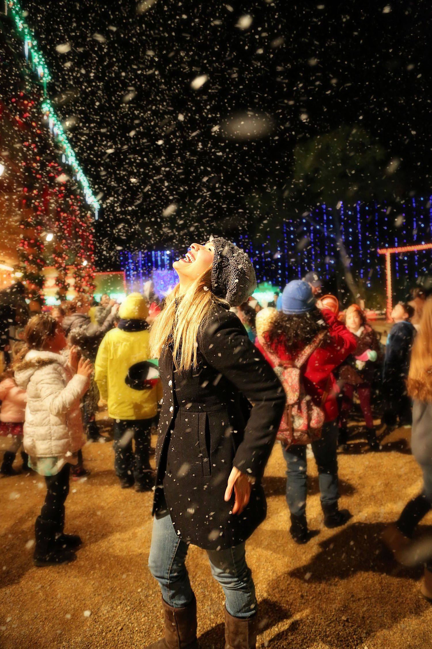 A woman in a beanie and black coat tilts her face skyward as artificial snow falls around crowds enjoying a night holiday lights festival.