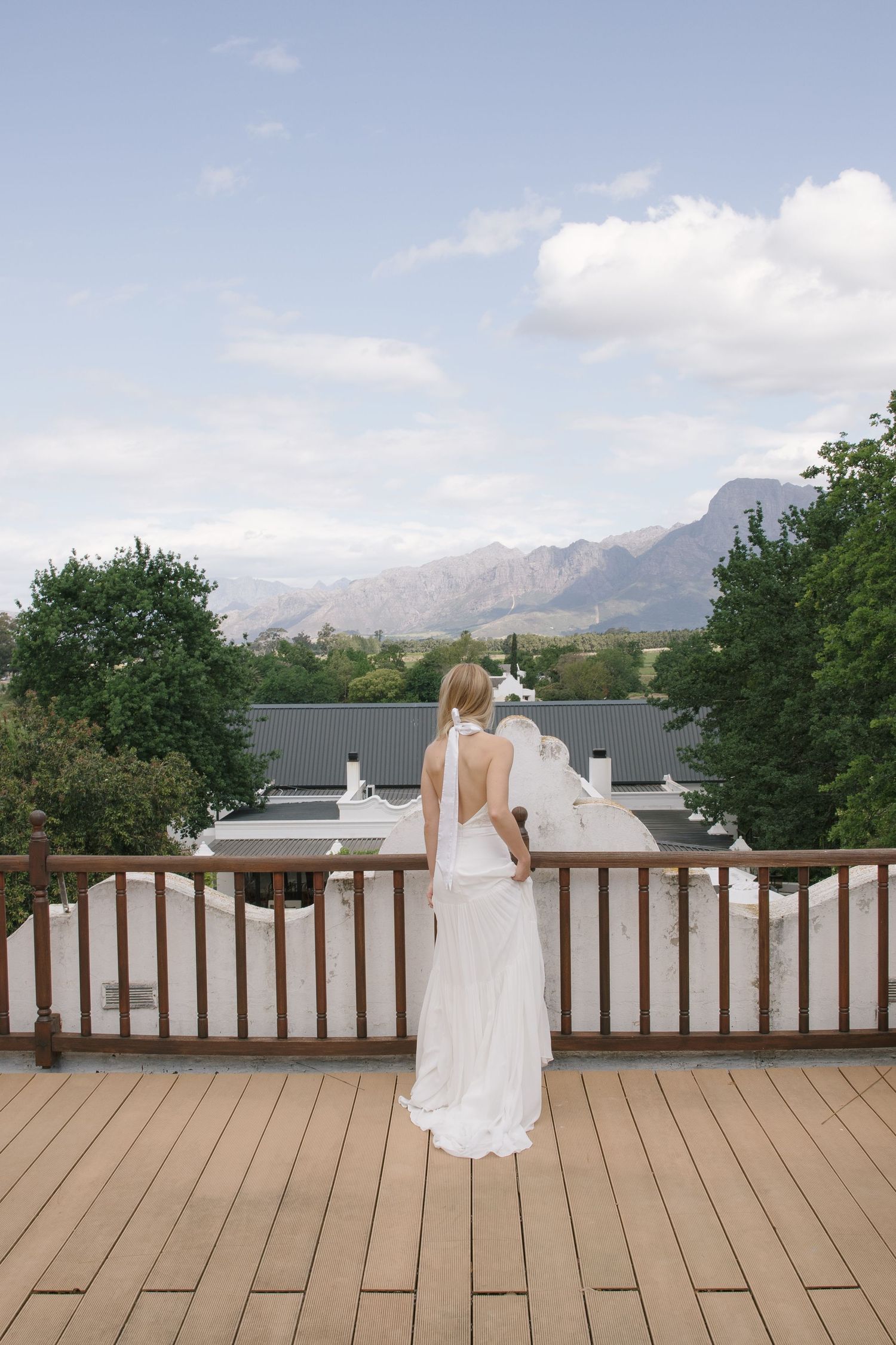 Bride in white backless gown stands on wooden deck overlooking mountain landscape and green trees.
