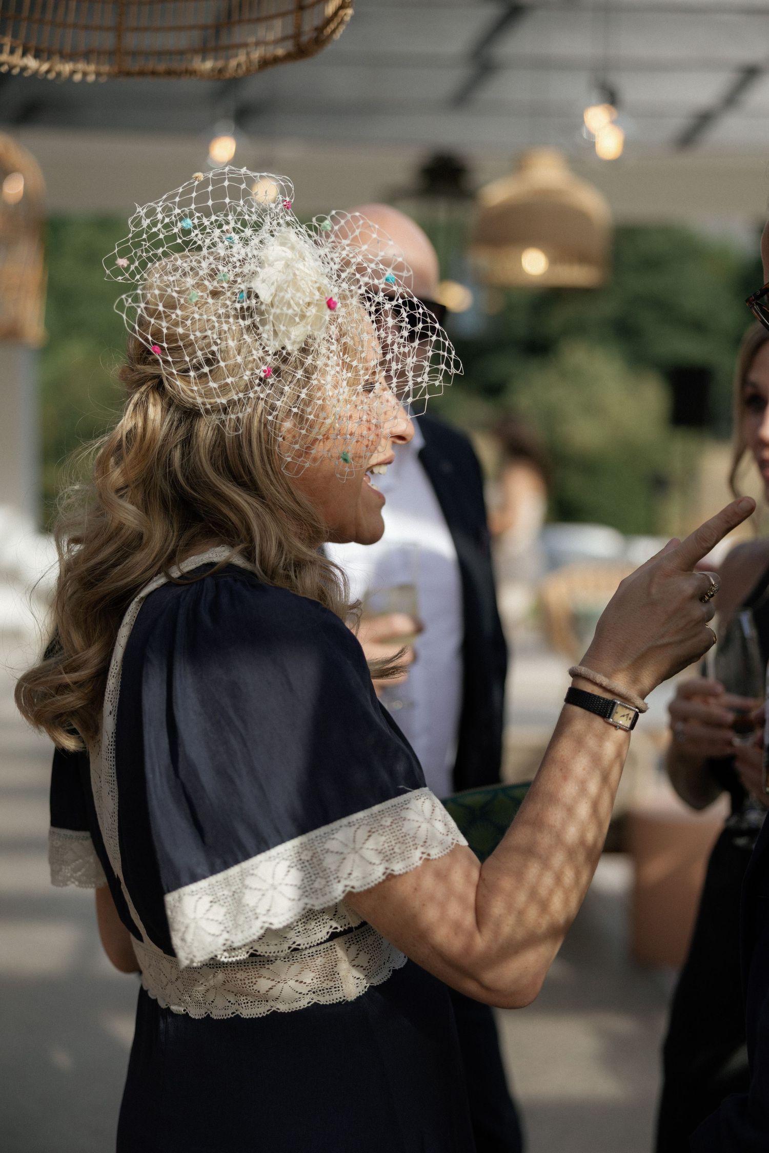 Woman in navy dress with lace trim and decorative birdcage veil fascinator at an outdoor event.