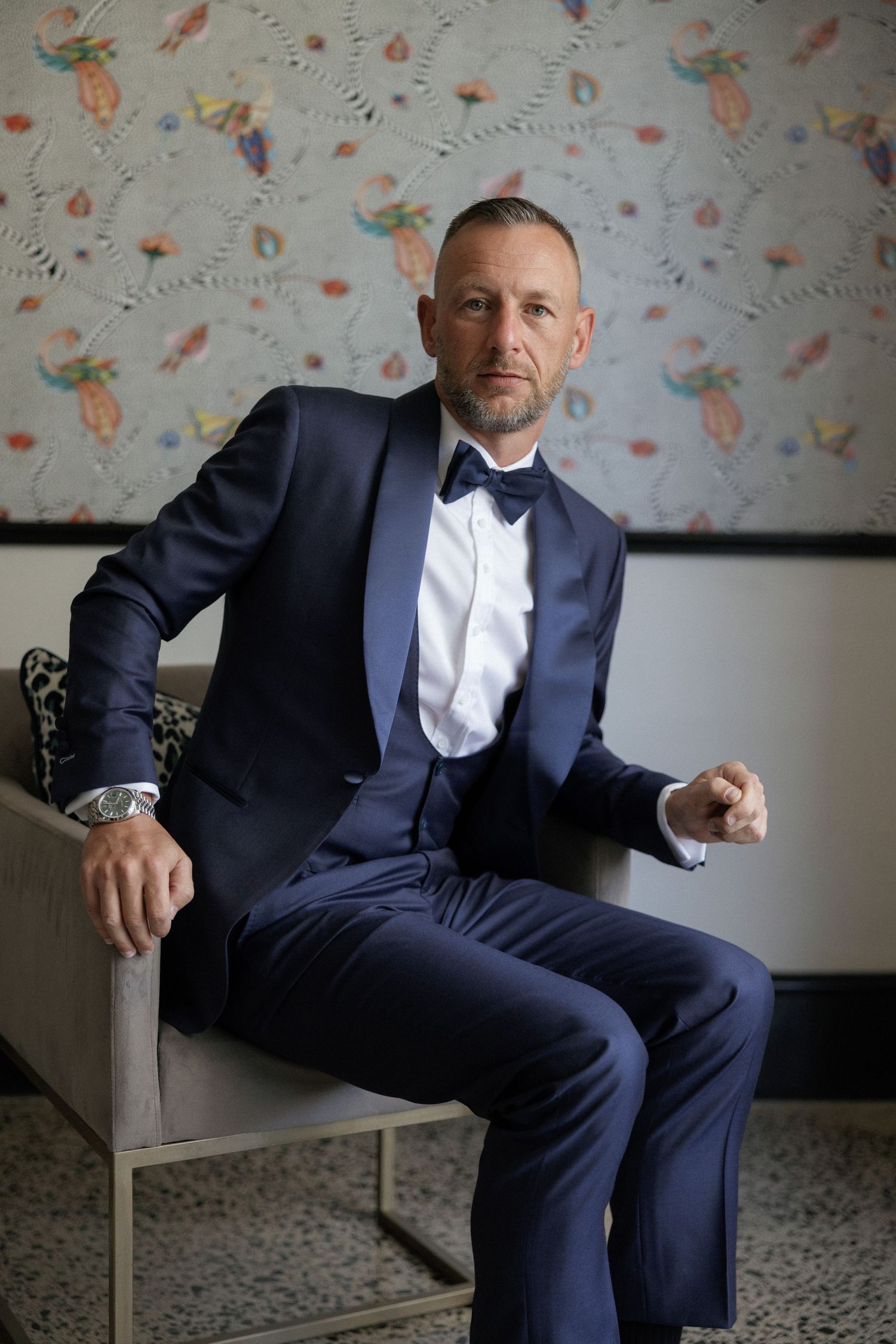 Man in navy blue tuxedo with bow tie seated confidently in chair against decorative wallpaper background.