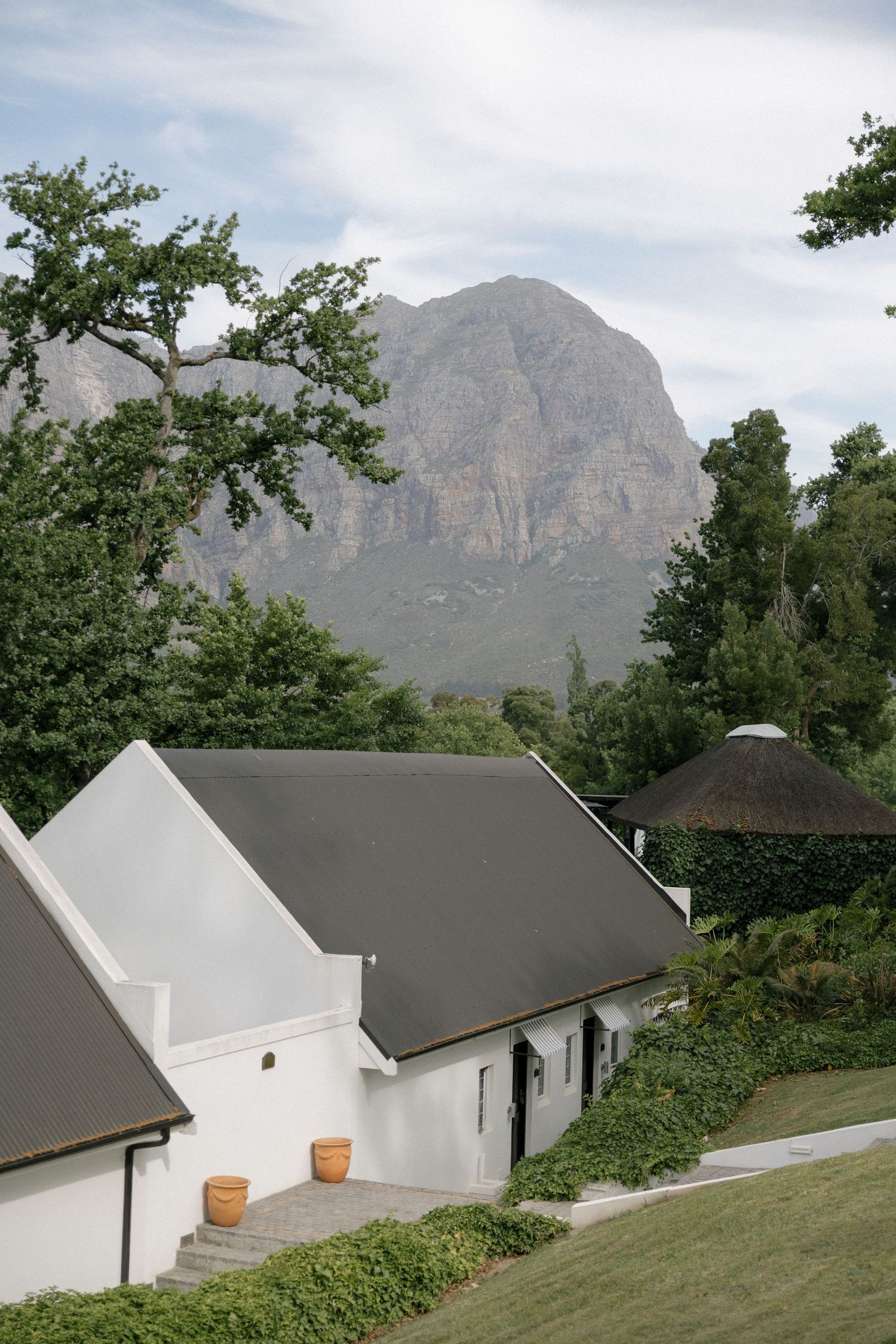White Cape Dutch buildings with dark roofs set against lush greenery and a dramatic rocky mountain backdrop under cloudy skies.