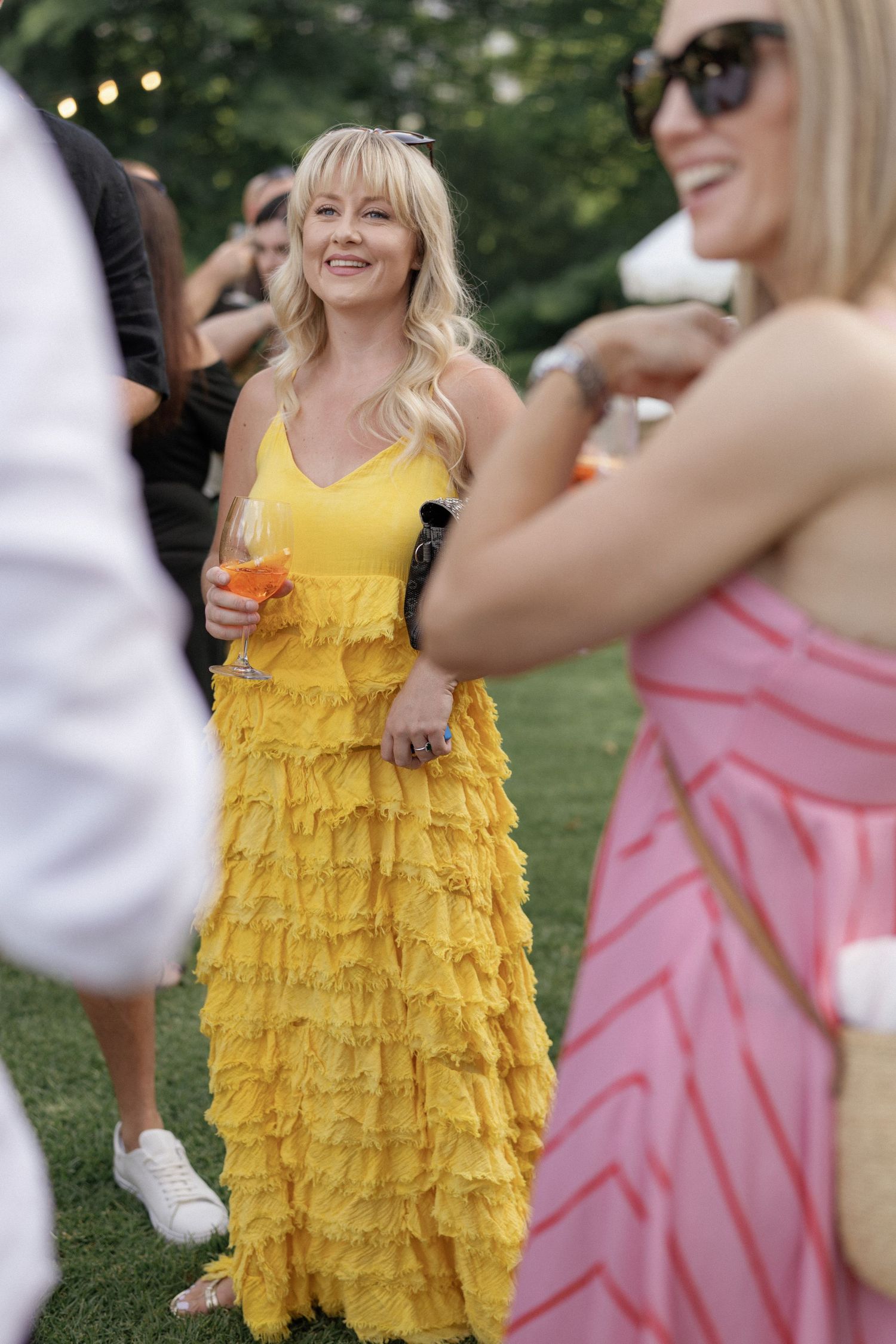Woman in yellow ruffled maxi dress holding a cocktail at an outdoor garden party event.