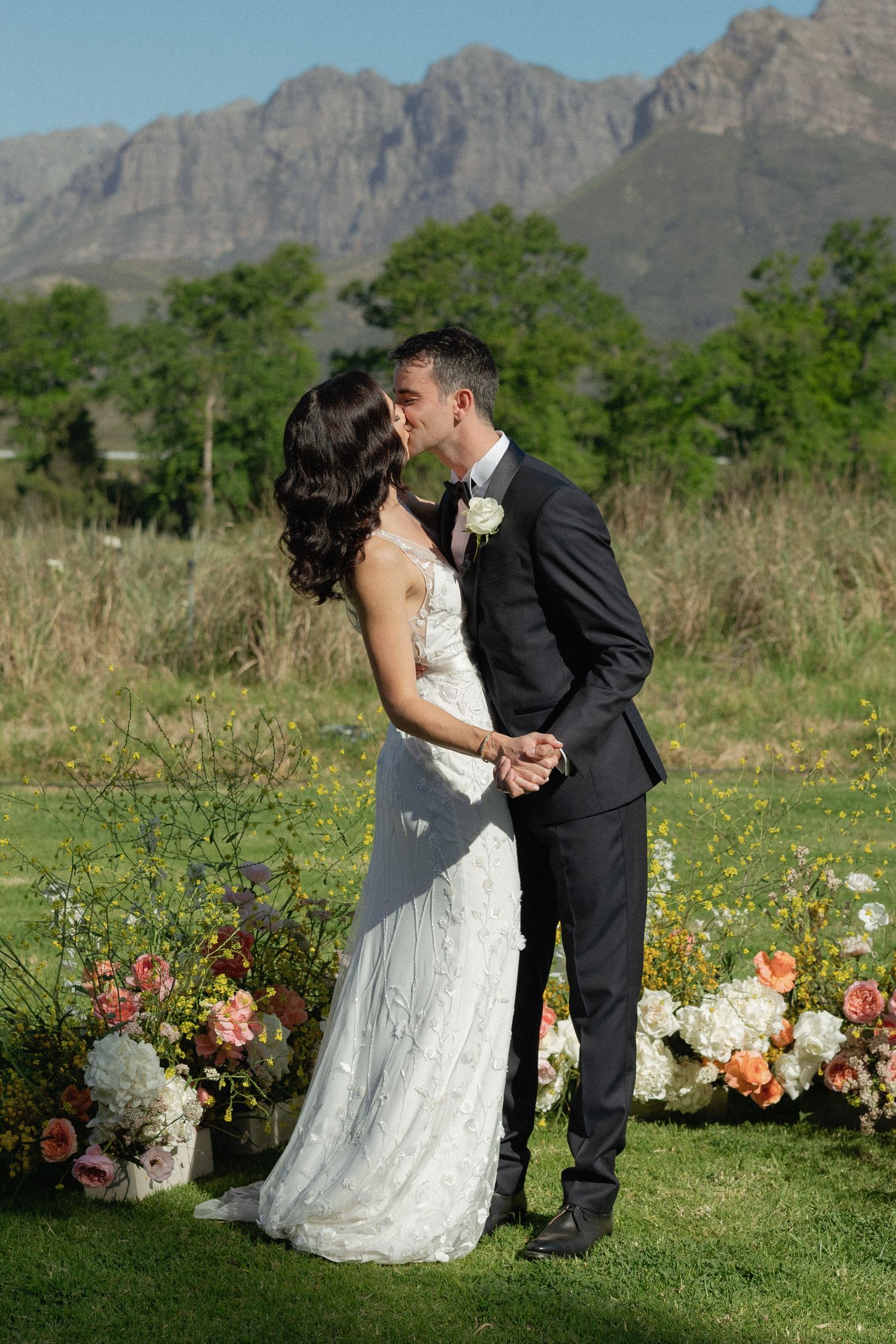 Bride and groom sharing first kiss at outdoor wedding altar surrounded by colorful floral arrangements and mountains.