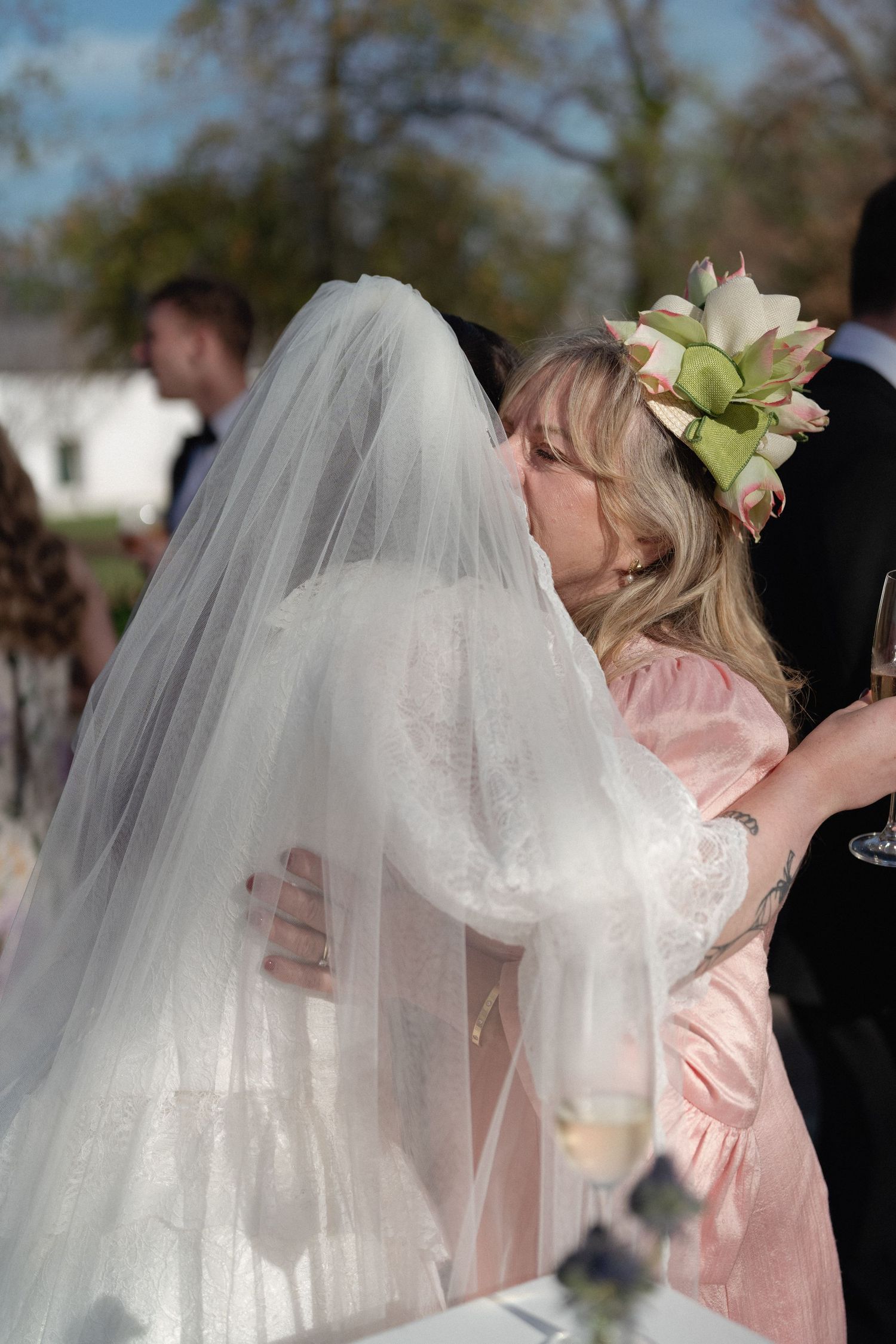 Bride in white veil embracing a woman in pink dress with floral headpiece at an outdoor wedding reception.