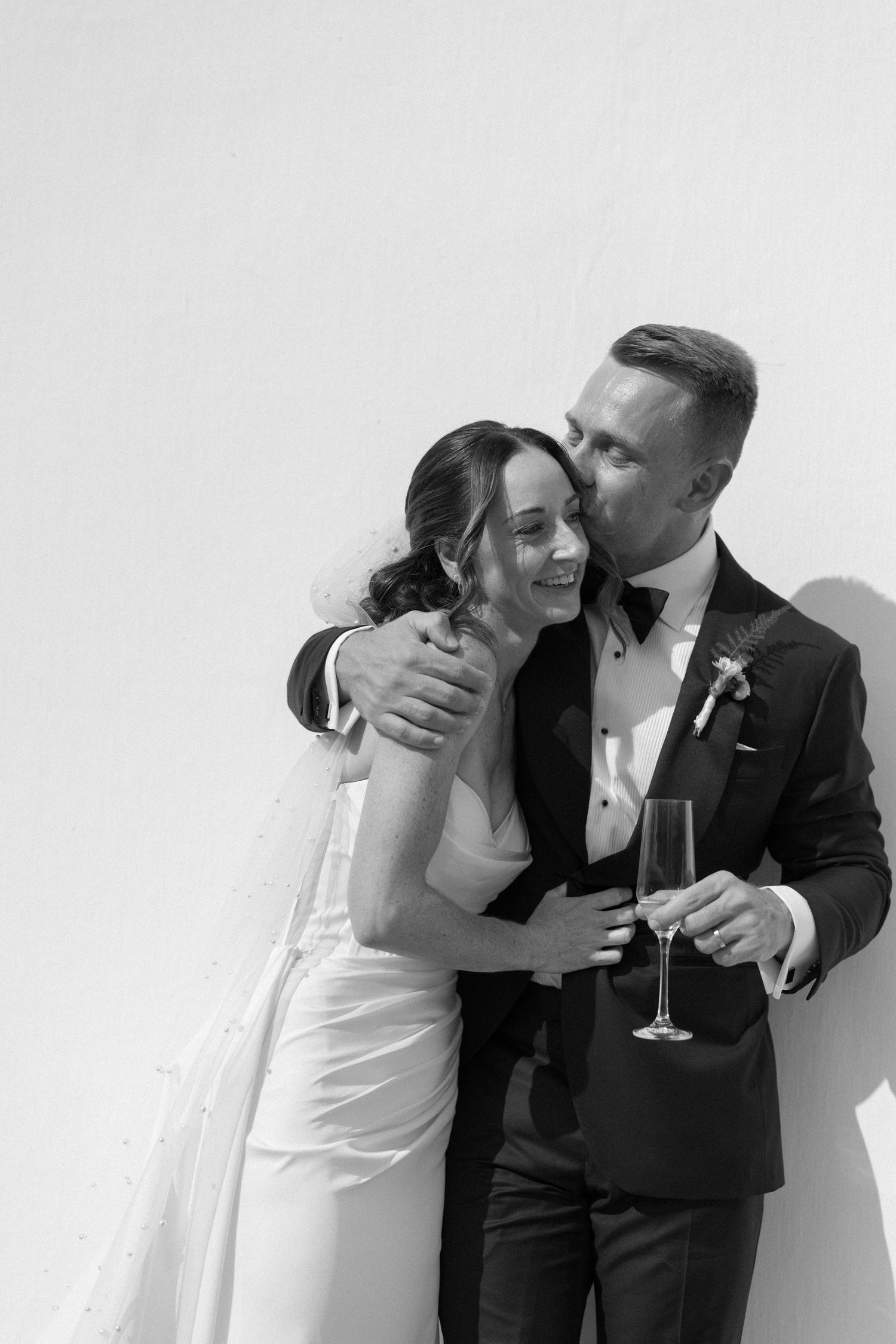 Black and white photo of a groom kissing bride on cheek while holding champagne flute against white wall.