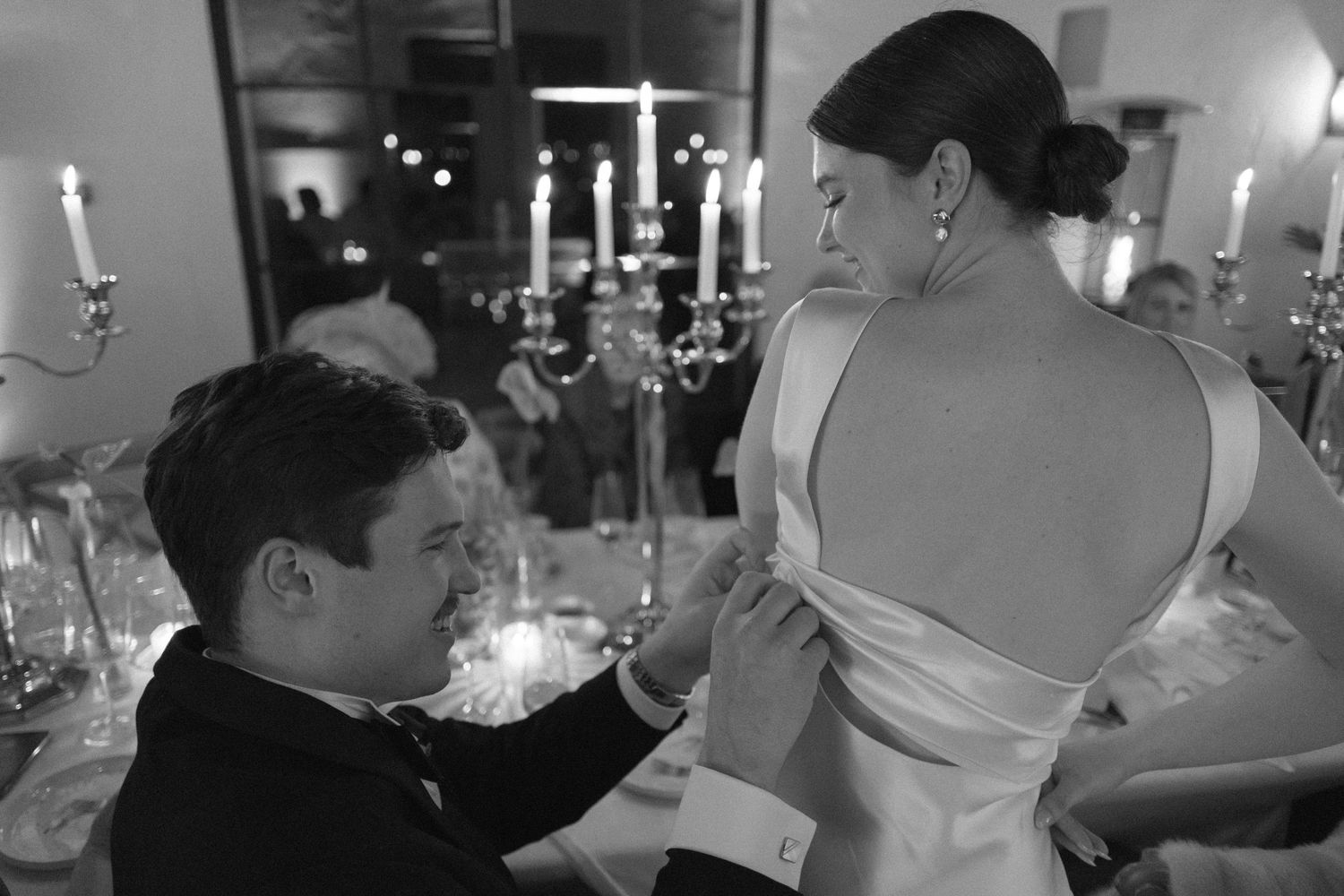 Black and white photo of groom fastening bride's wedding dress at elegant candlelit reception.