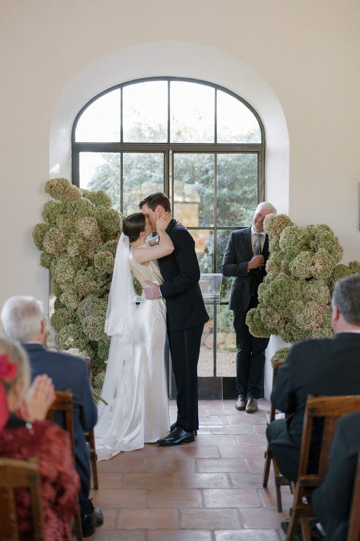 Bride and groom kiss at altar flanked by hydrangea arrangements before seated wedding guests.