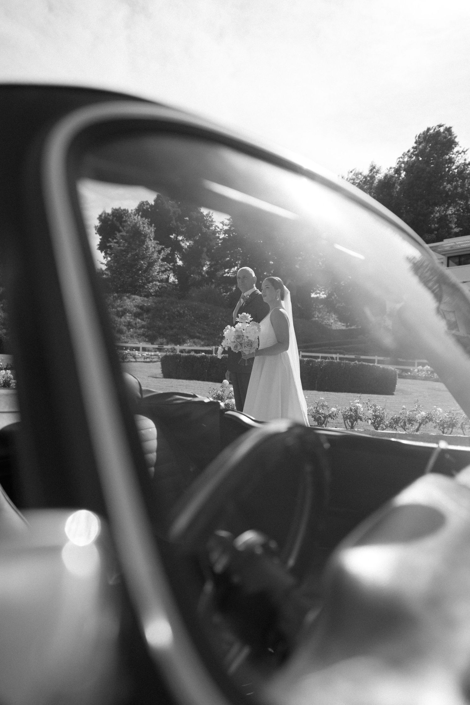 Black and white wedding photo of bride and groom kissing, reflected in a vintage car window outdoors.