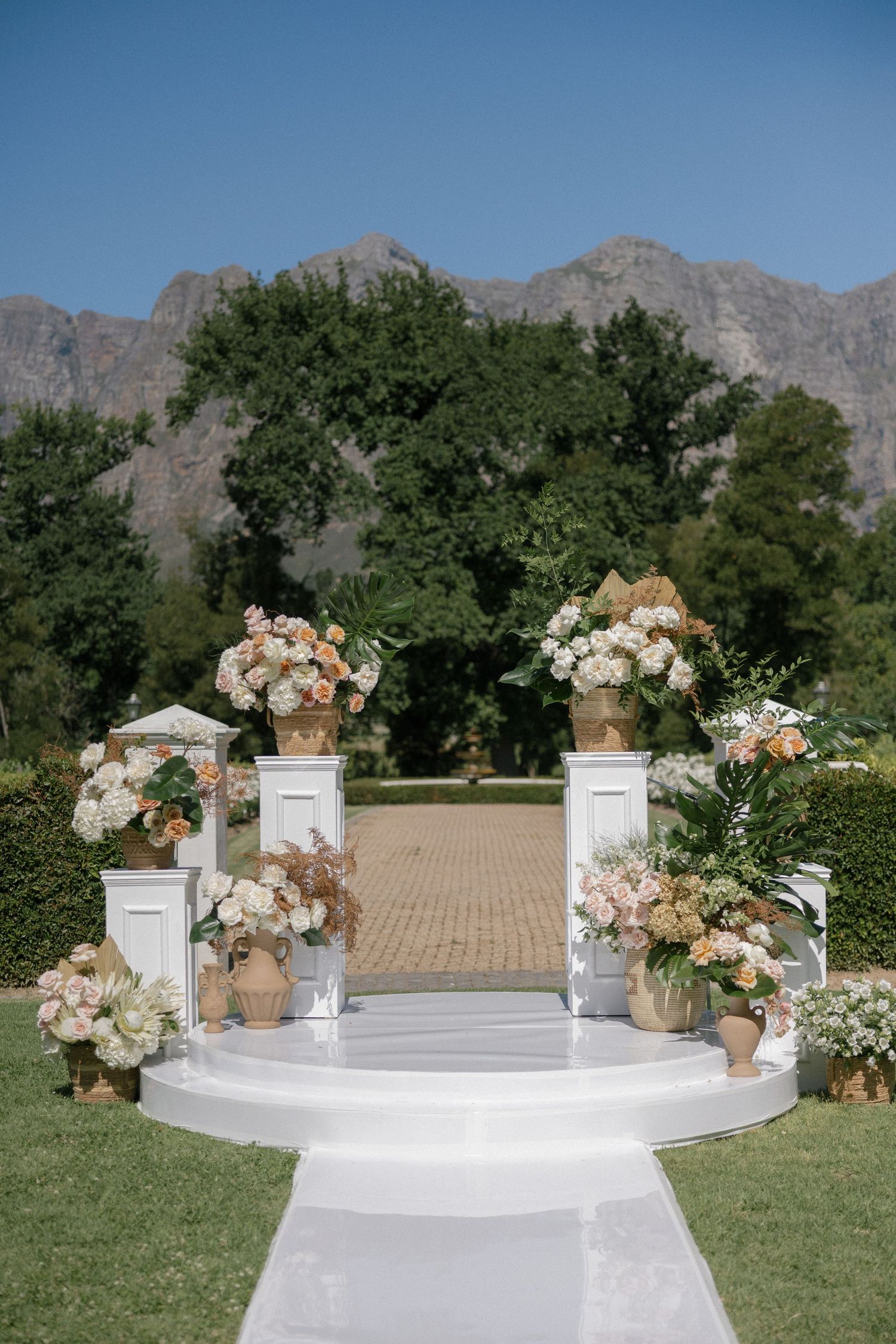Elegant outdoor wedding ceremony setup with white pillars, floral arrangements, and mountain backdrop on a sunny day.