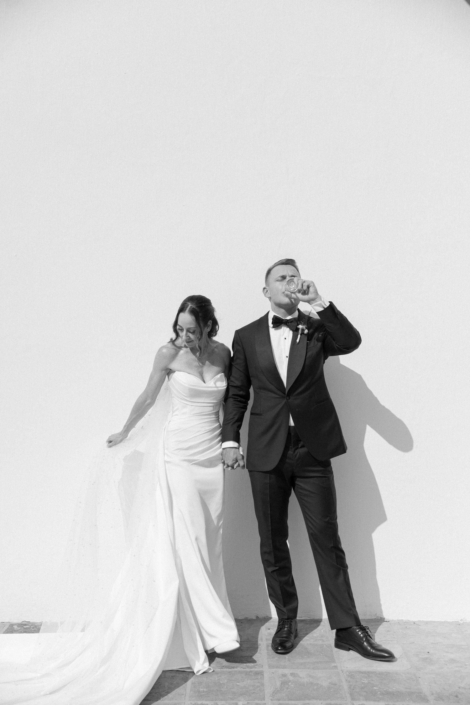 Bride and groom in black and white photo, holding hands against white wall, groom drinking champagne.