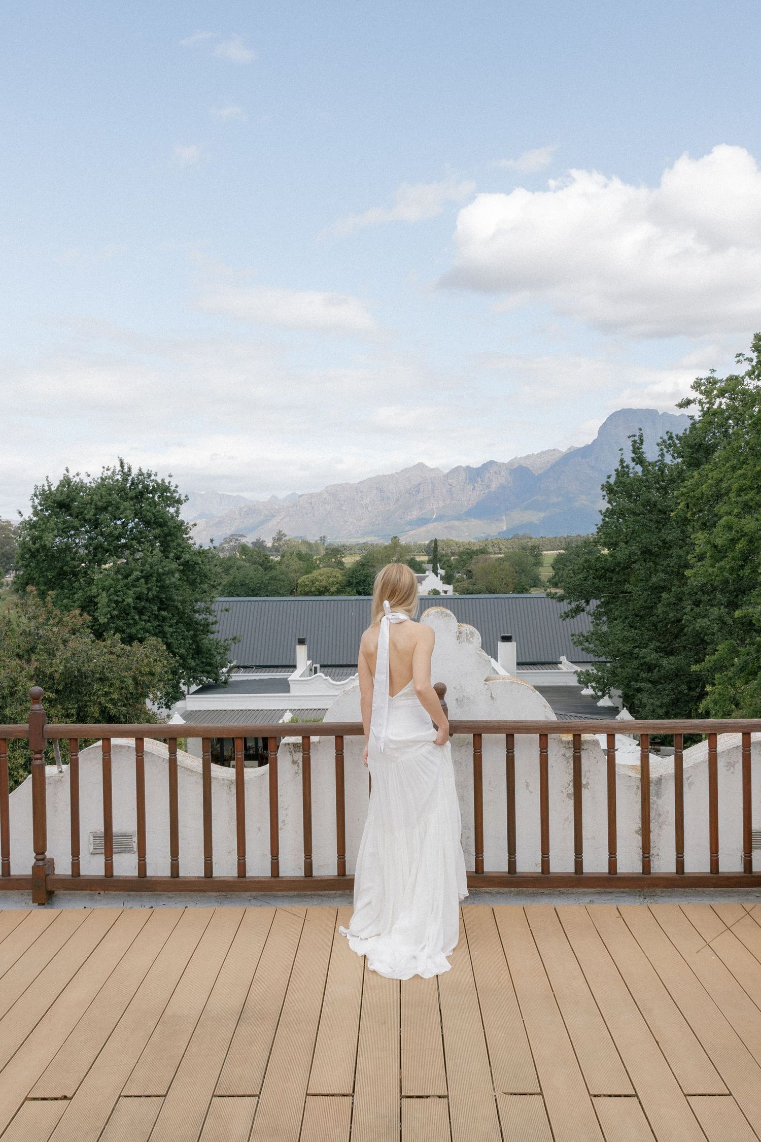 Bride in backless white gown stands on wooden deck overlooking mountain landscape.