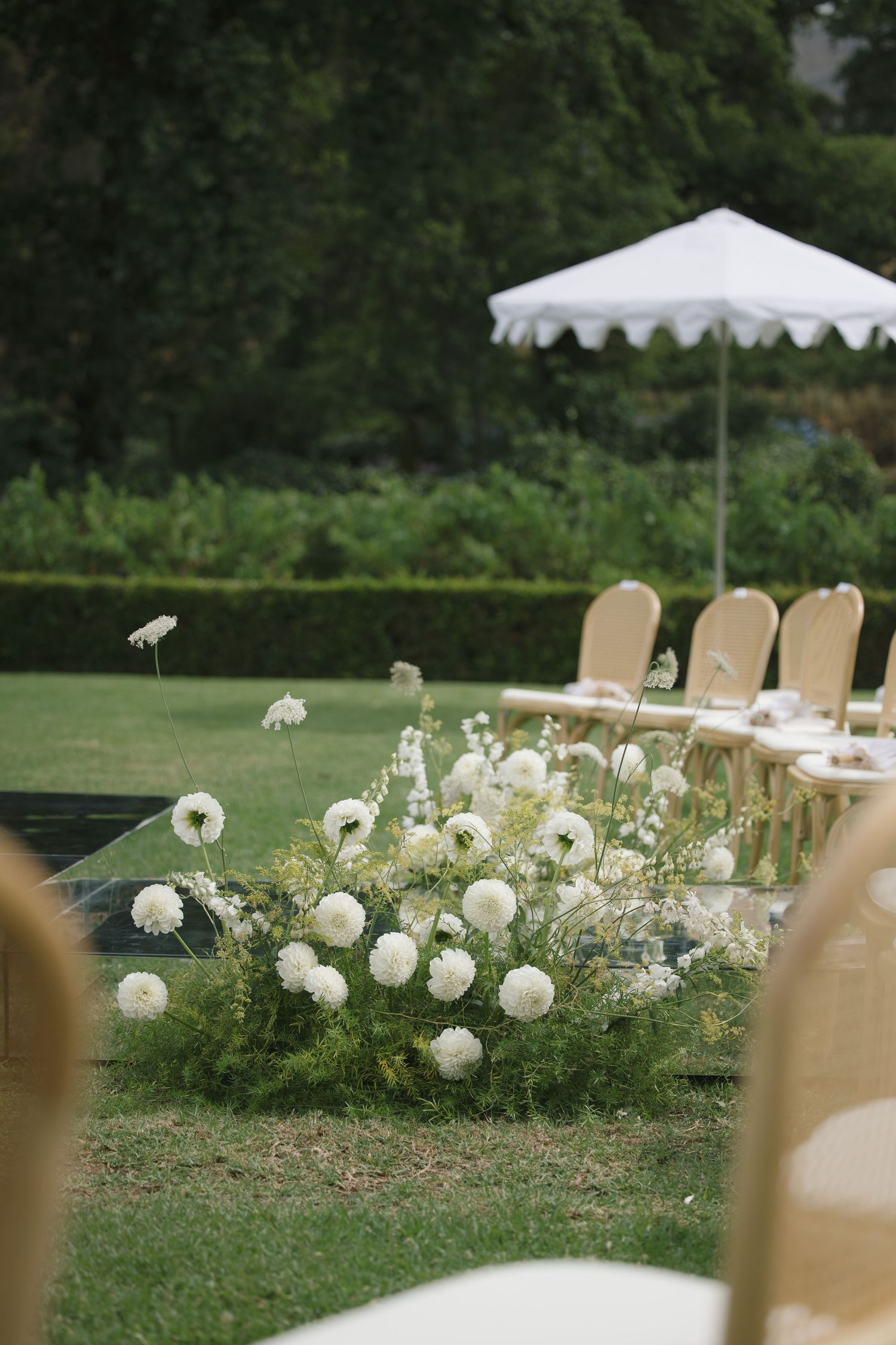 Elegant outdoor wedding setup with white pompom flowers, gold chairs, and a white scalloped umbrella on a lush green lawn.
