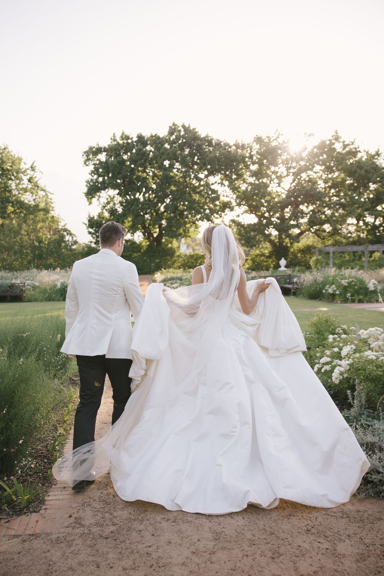 Bride and groom walking hand in hand through a sunlit garden, bride in a flowing white gown with veil and groom in white jacket.