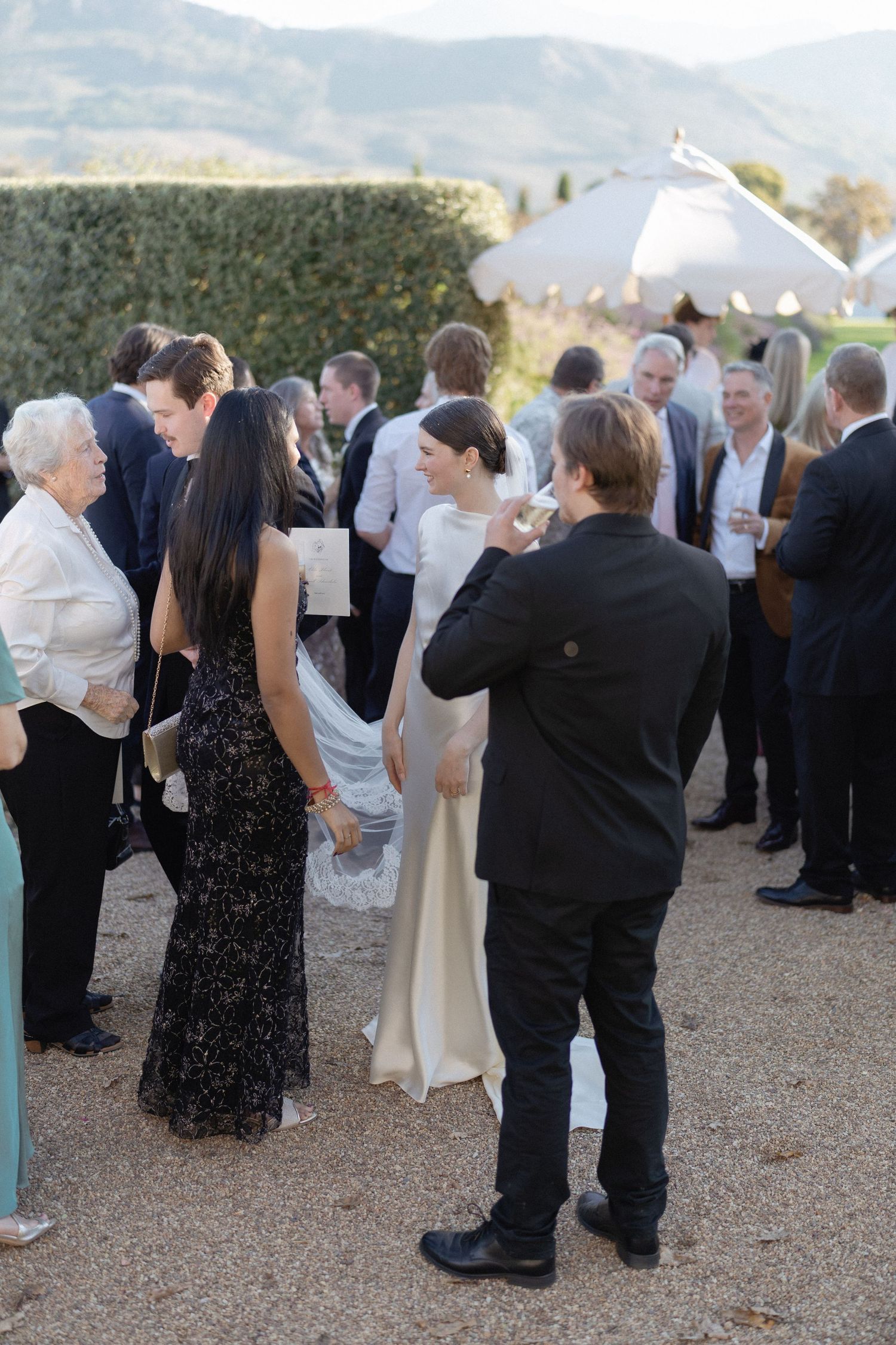 Bride in ivory gown mingles with wedding guests during outdoor cocktail hour with mountain views and hedge backdrop.