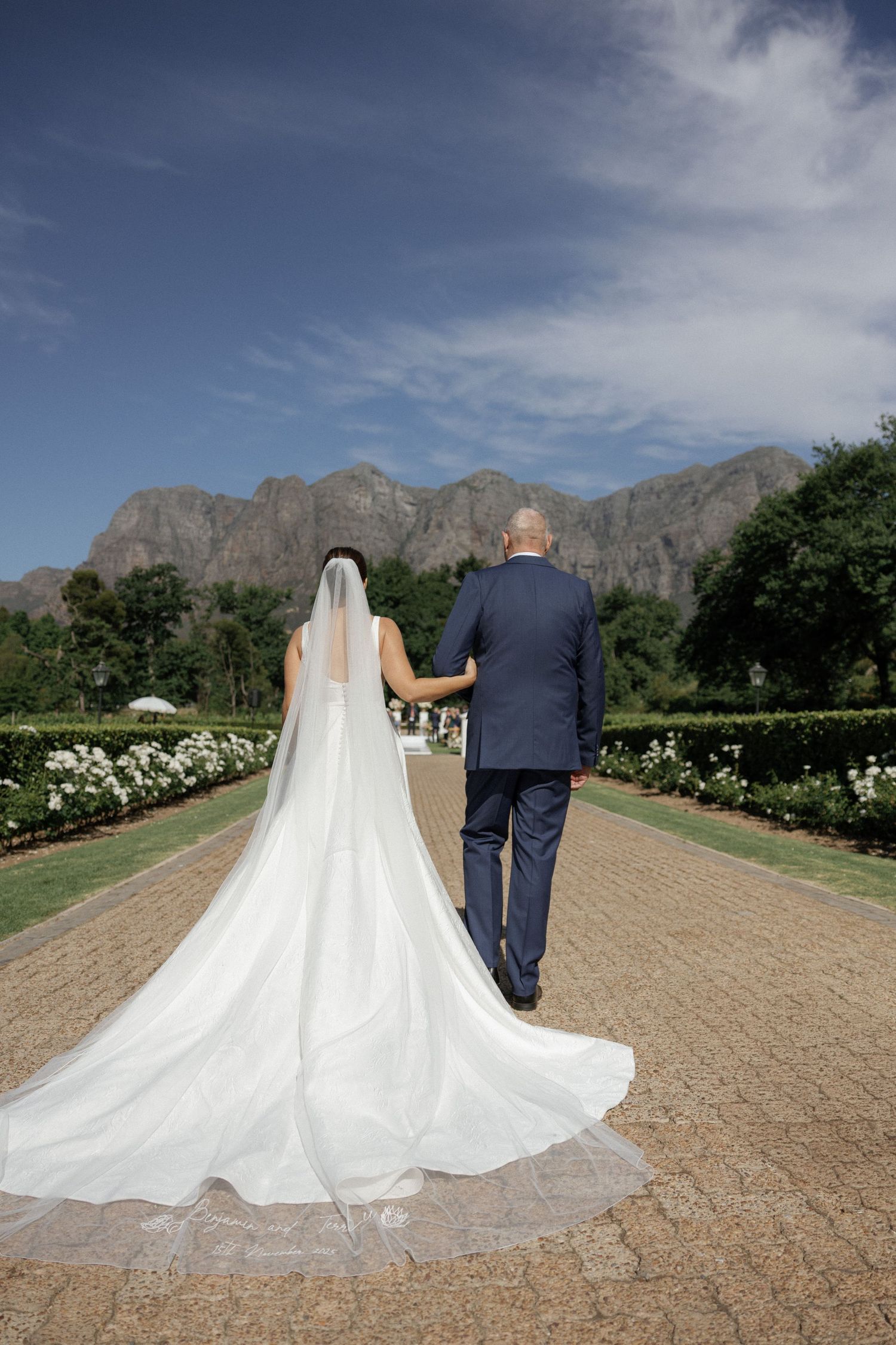 Bride and groom walking hand in hand down a garden path with mountain views on their wedding day.