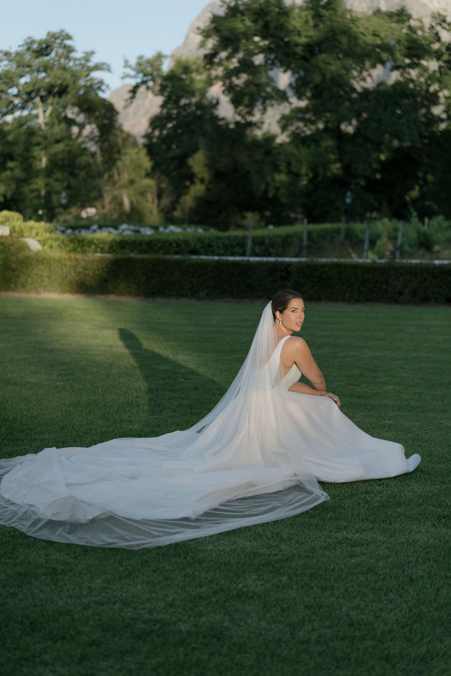 Bride in white gown and long veil sits gracefully on green lawn with mountain backdrop.