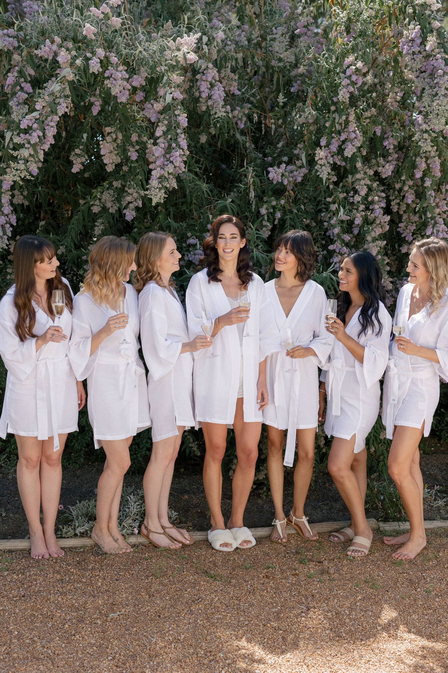 Seven bridesmaids in white robes holding champagne glasses, laughing together in front of a blooming floral wall.