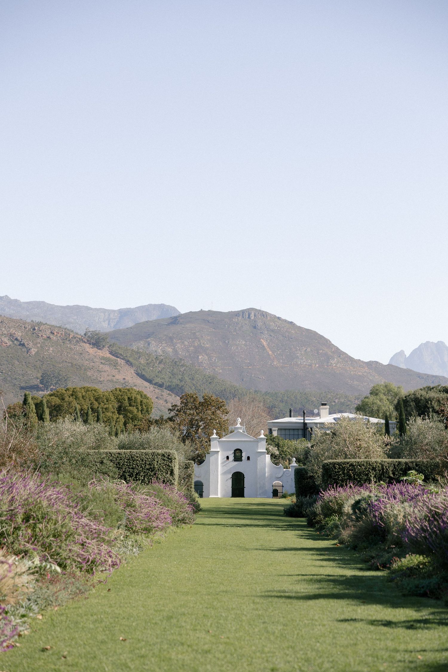 White Cape Dutch chapel at end of lush garden path with mountain backdrop in Franschhoek, South Africa.