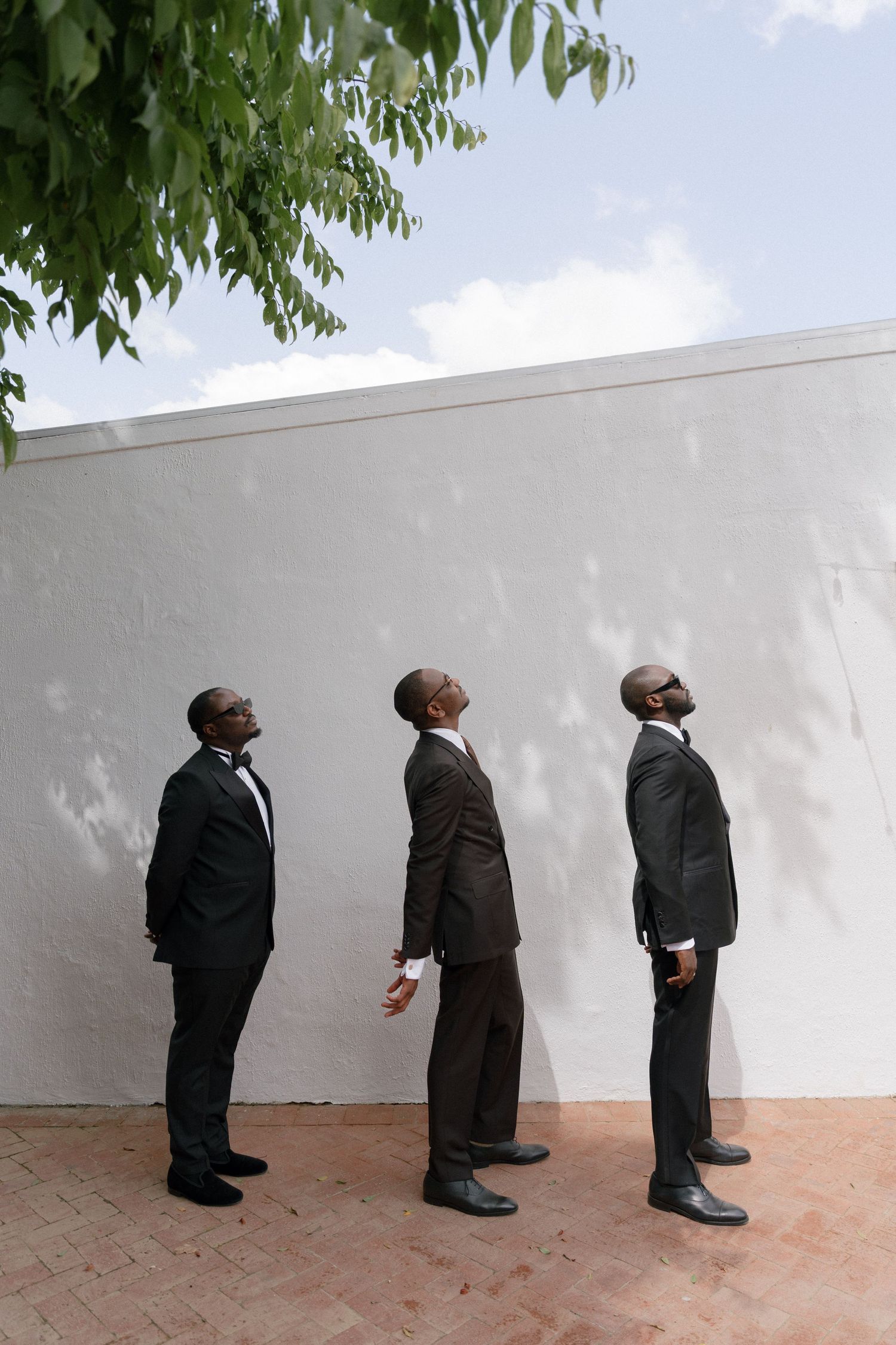 groom & groomsmen in dark suits and bow ties stand against a white wall, all looking upward toward the sky.